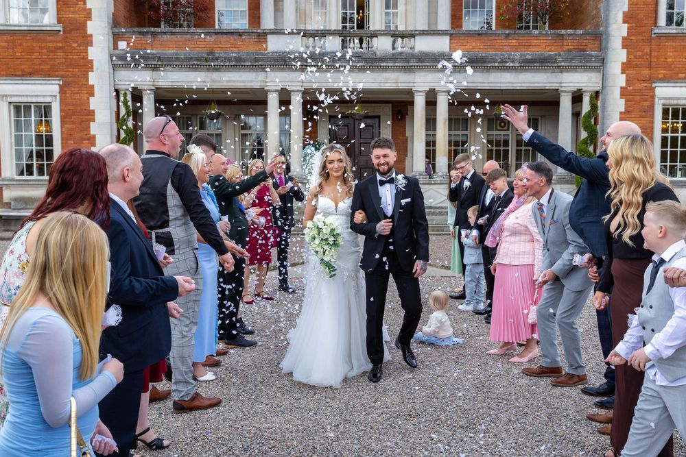 Bride and groom walk through confetti shower outside elegant brick manor surrounded by smiling wedding guests.