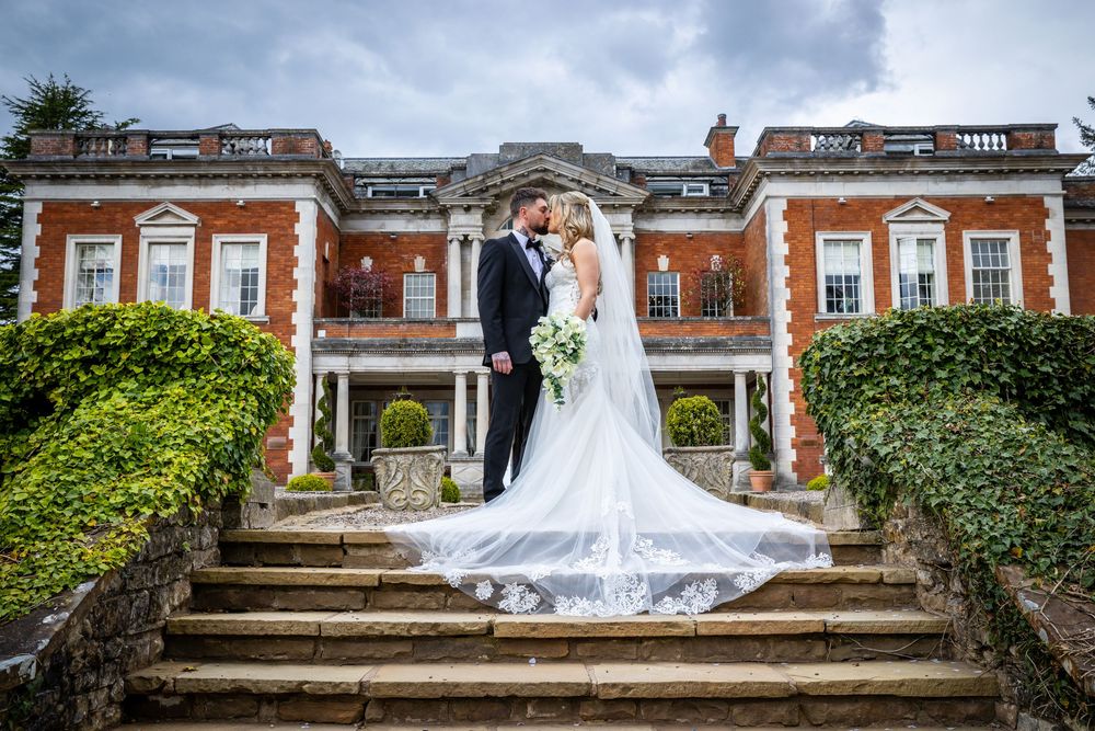 Bride and groom kissing on stone steps of elegant red brick mansion wedding venue with flowing veil.