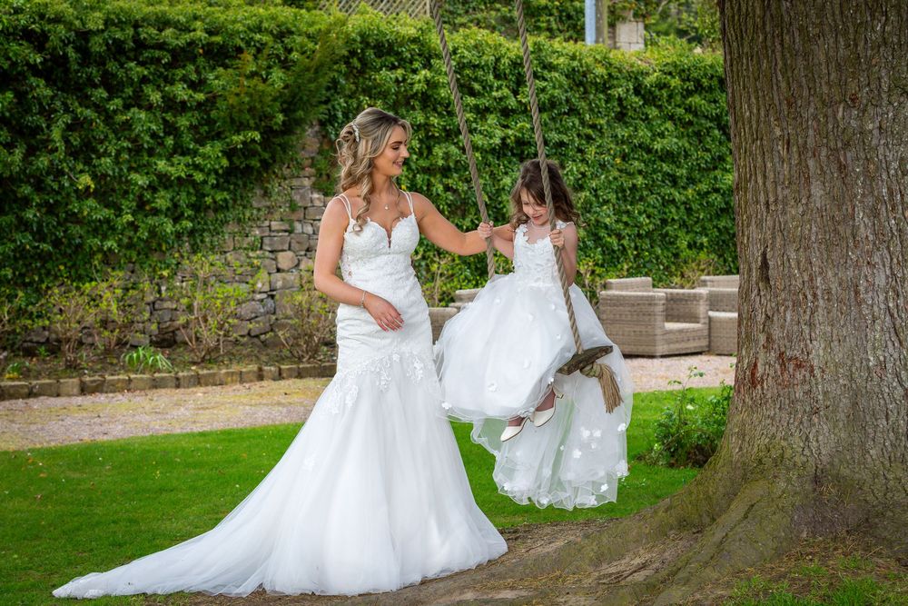 Bride in white mermaid gown pushes flower girl on tree swing in lush garden wedding setting.