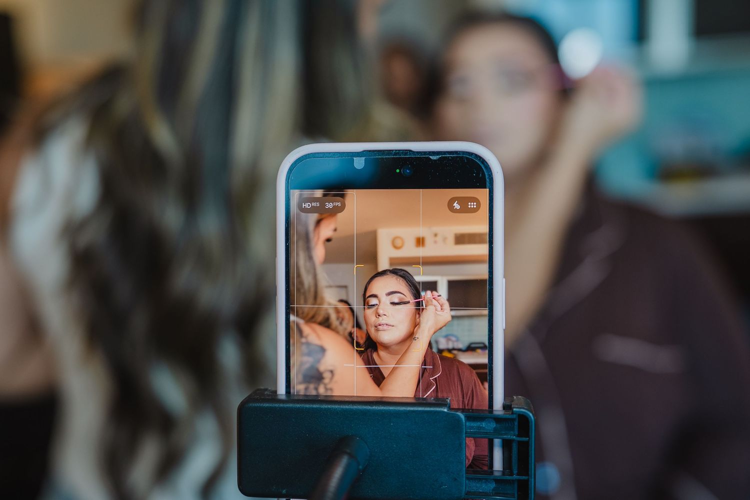 A smartphone on a tripod records a woman applying makeup, capturing her reflection on screen in a beauty studio.