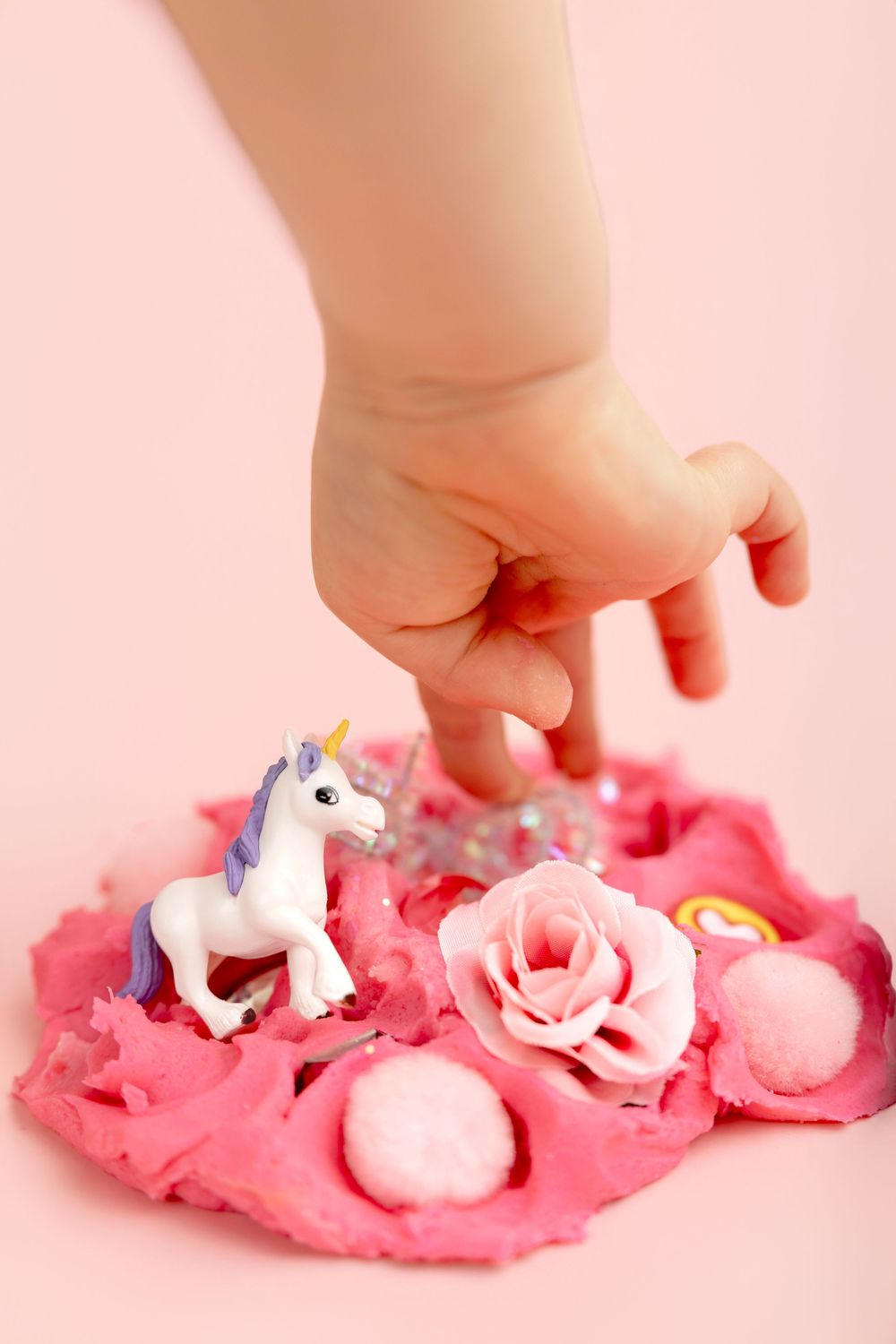 A child's hand reaching into pink sensory play dough with a unicorn figurine, rose, pom poms, and gems.