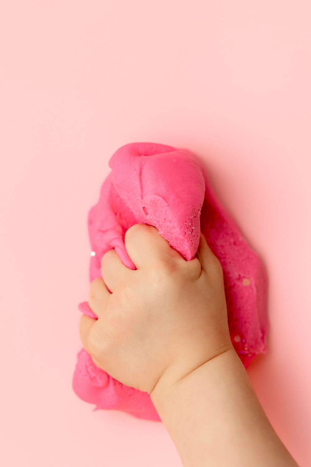 A child's hand squeezing pink slime or play dough against a pink background.