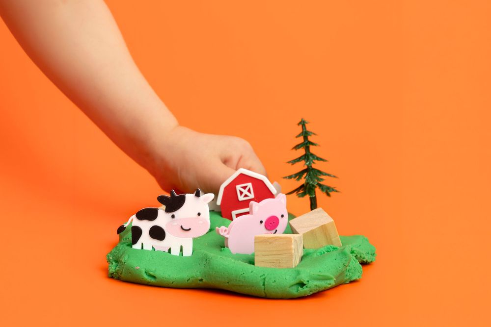 Child's hand placing a red barn toy on green play dough farm scene with cow, pig, and tree figures.