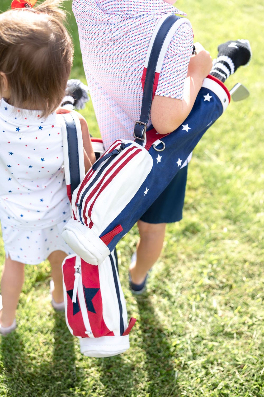 Adult carrying patriotic stars and stripes golf bag with child in matching American flag outfit on green grass.