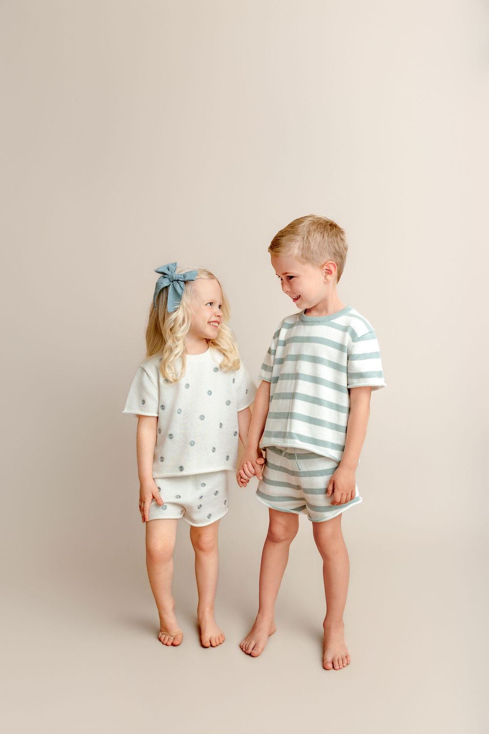 Two young children in matching pastel pajama sets, a girl in dotted shorts and a boy in striped shorts, smiling at each other.