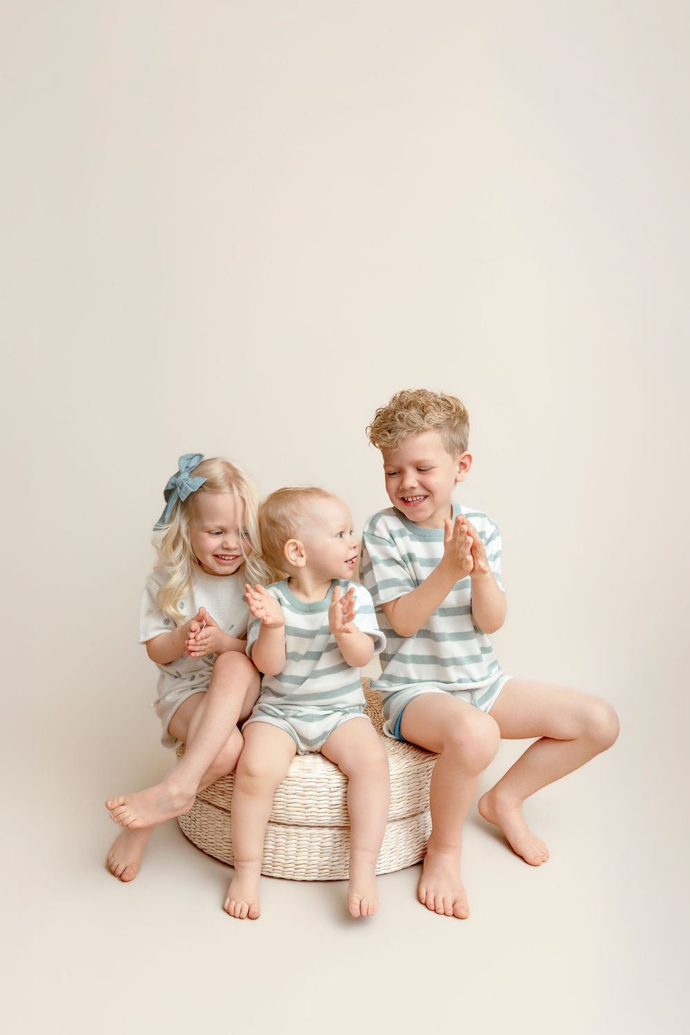 Three young siblings in matching striped outfits sit together on a woven pouf, laughing and clapping in a bright studio.