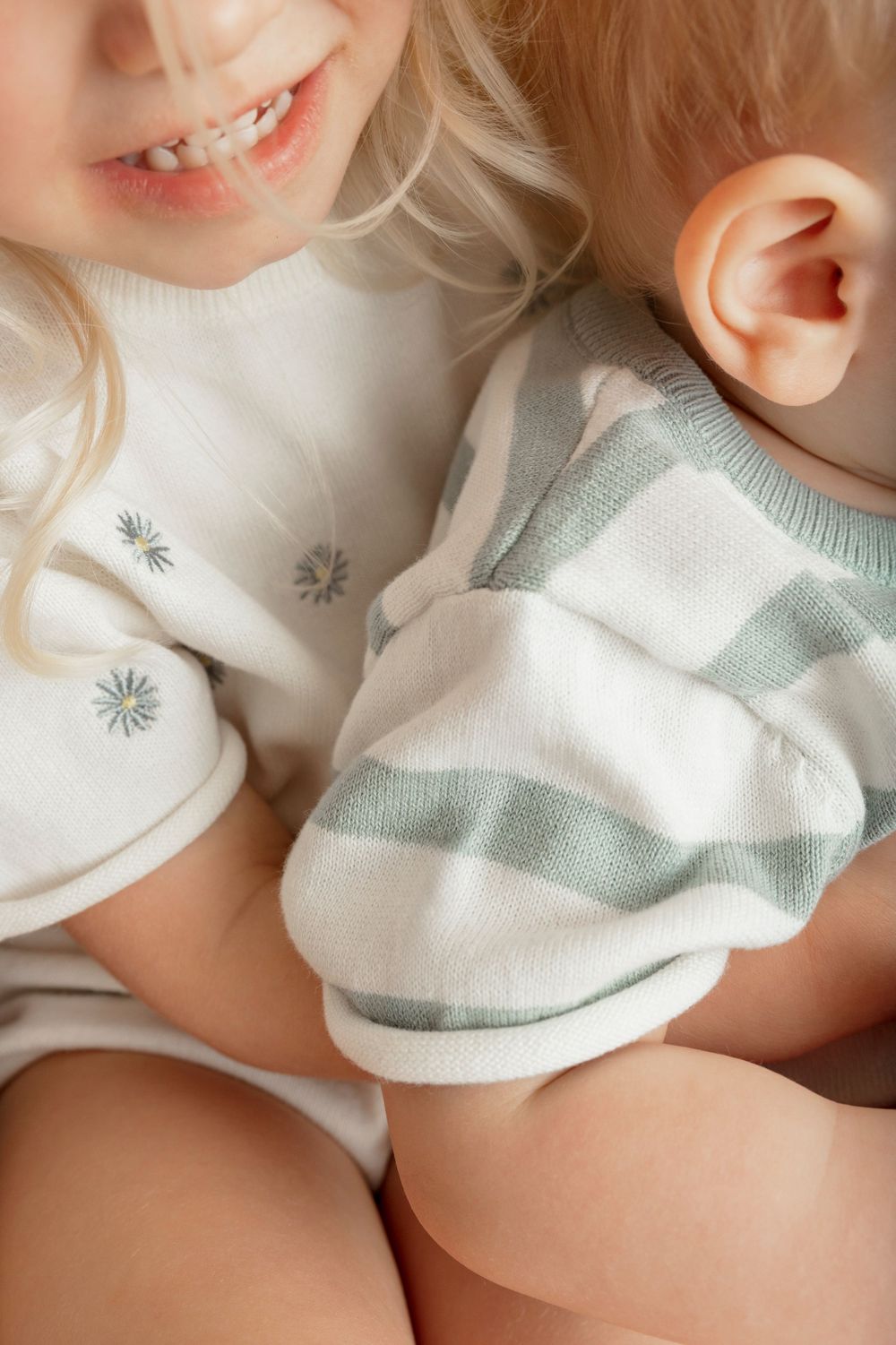 Two young children in soft pastel outfits, one smiling blonde girl hugging a baby in a striped romper.