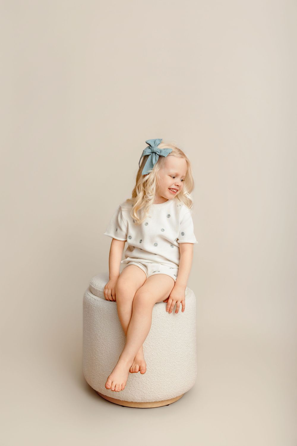 Young blonde girl in white polka dot outfit sitting on a round ottoman with a blue bow in her hair.
