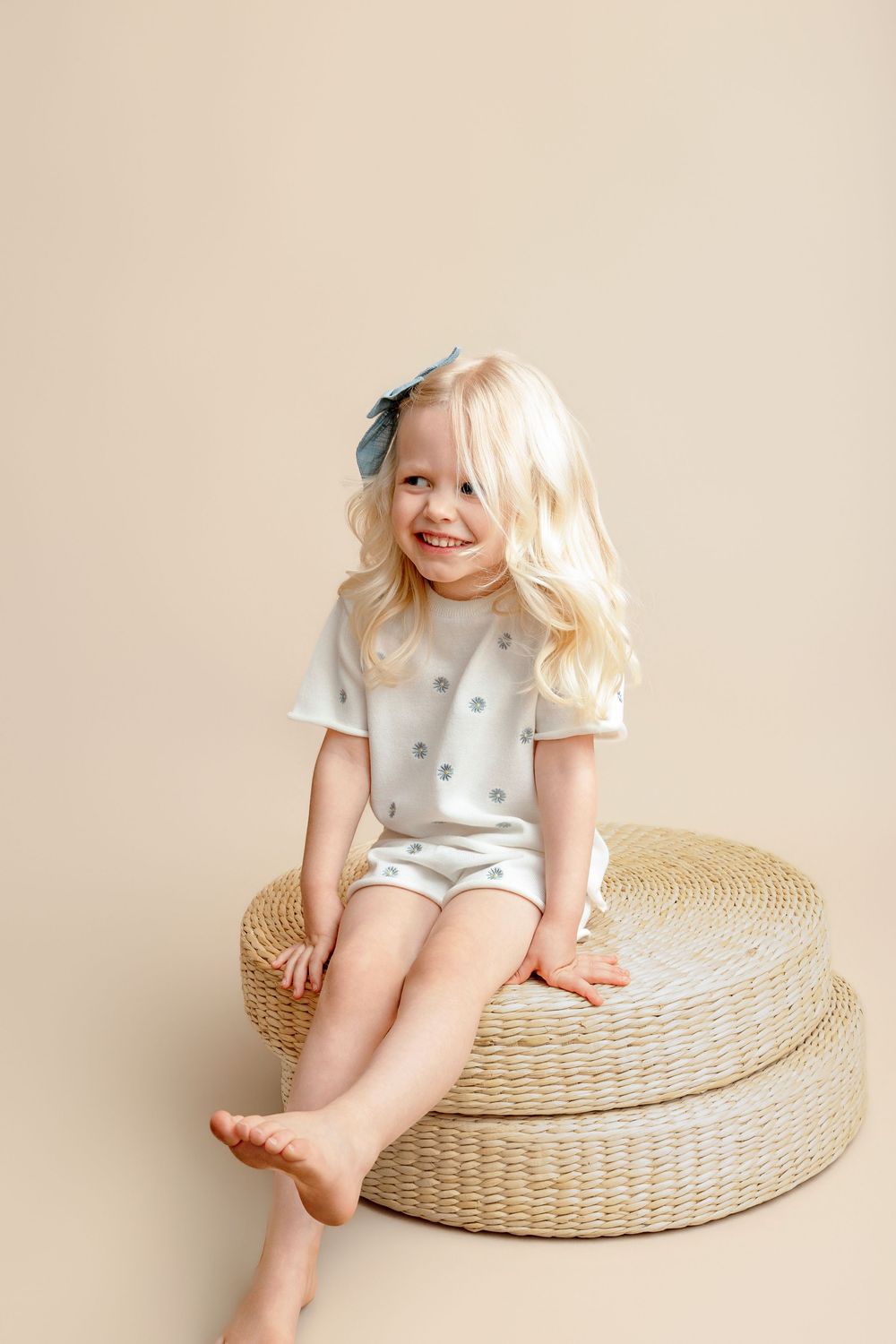 Smiling blonde toddler girl sitting on wicker poufs wearing a light gray polka dot romper with a bow headband.