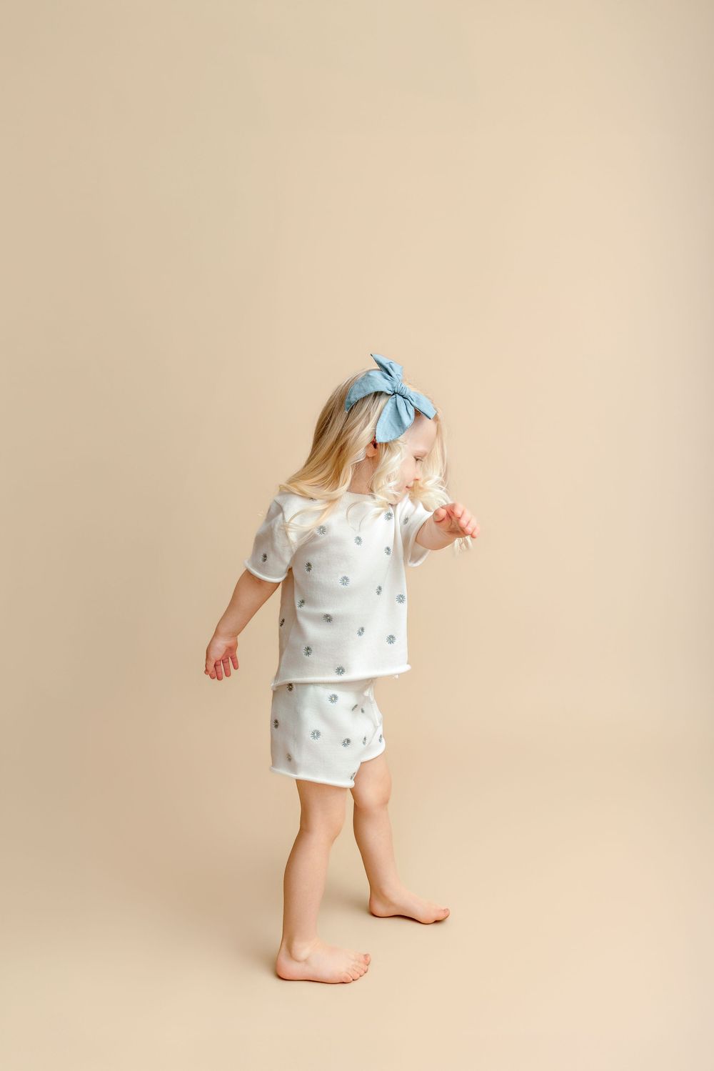 Toddler girl in white polka dot shorts set with blue bow headband, standing barefoot on beige background.