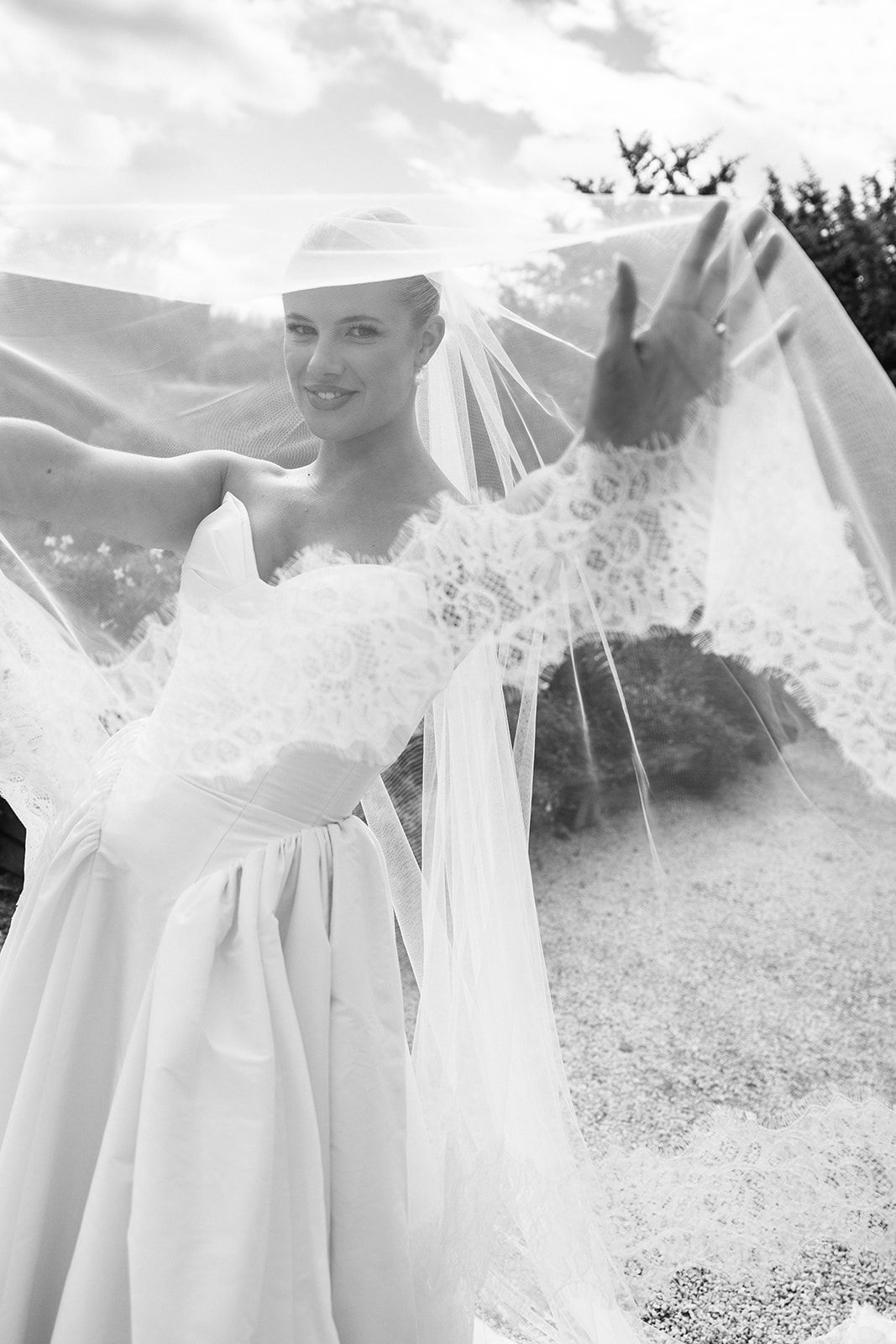 Black and white photo of a smiling bride in a white gown lifting her lace veil outdoors on her wedding day.