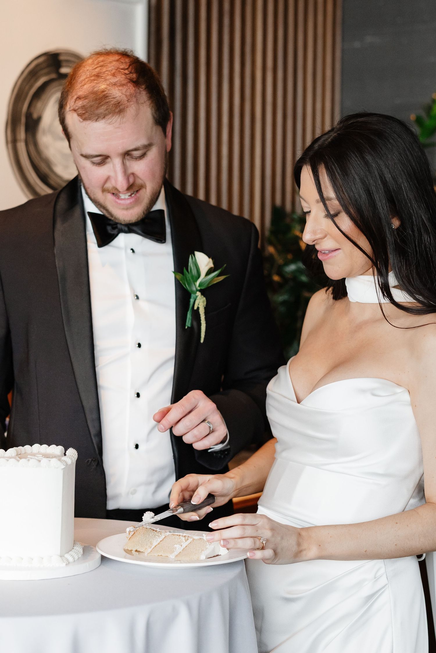 Bride and groom cutting their white wedding cake together, groom in tuxedo with boutonniere, bride in strapless gown.