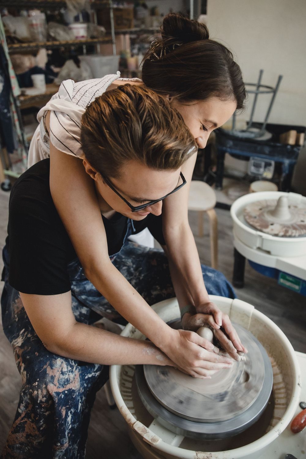 Pottery instructor teaching student to shape clay on a spinning wheel in a ceramic studio workshop.