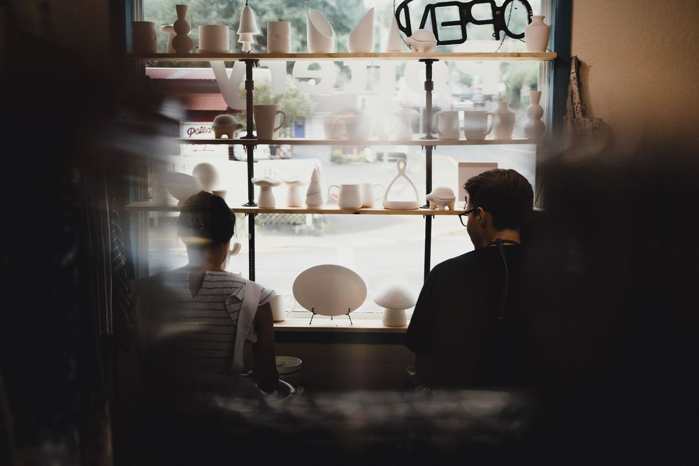Two people sit silhouetted at a cafe counter by a bright window surrounded by shelves with plants and ceramics.