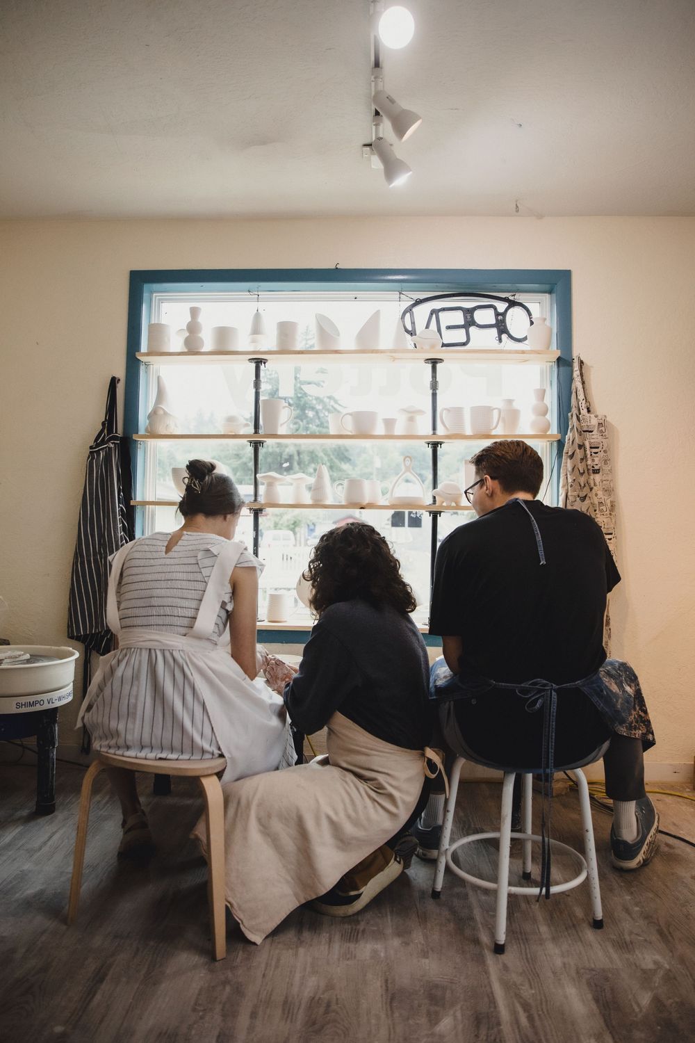 Three people sit with backs to camera in pottery studio, shelves of ceramic pieces displayed by window.