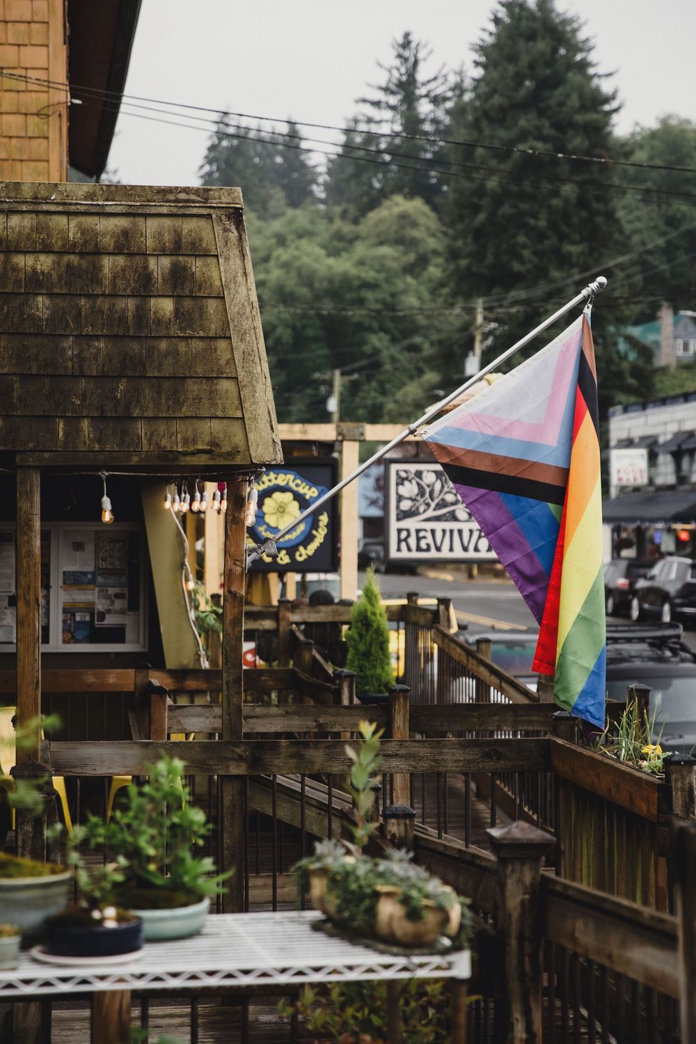Rainbow pride flag flying on wooden building deck with Revival sign and lush green trees in background.