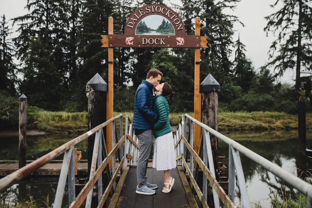 Couple kissing on a wooden dock bridge under a rustic sign surrounded by tall evergreen trees.