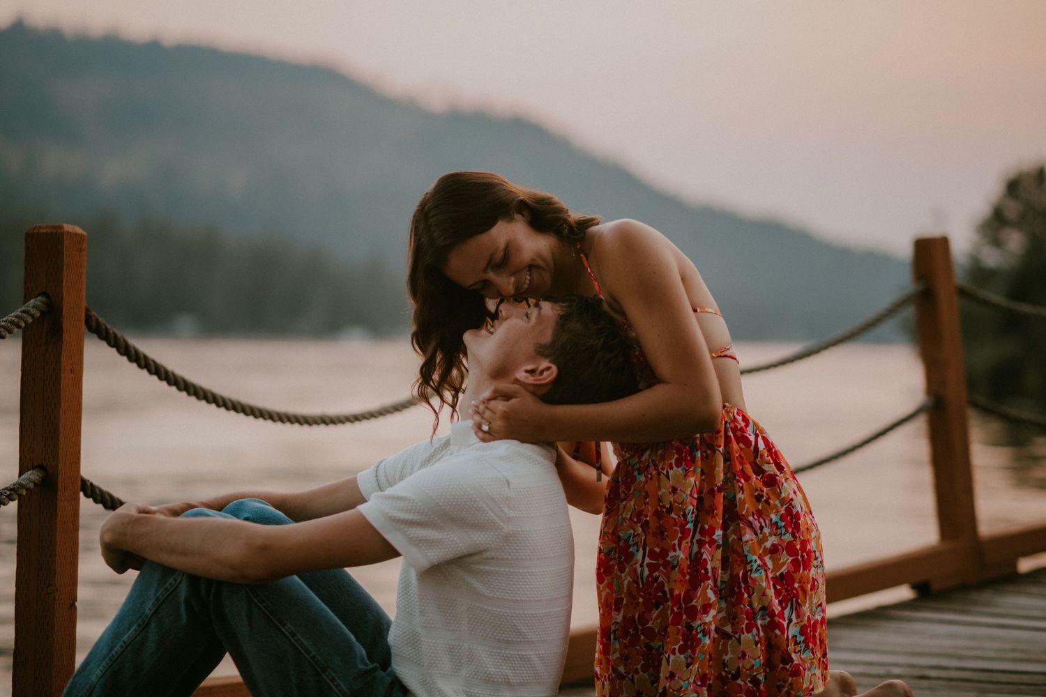 A couple shares a romantic moment on a wooden dock at sunset with misty mountains in the background.