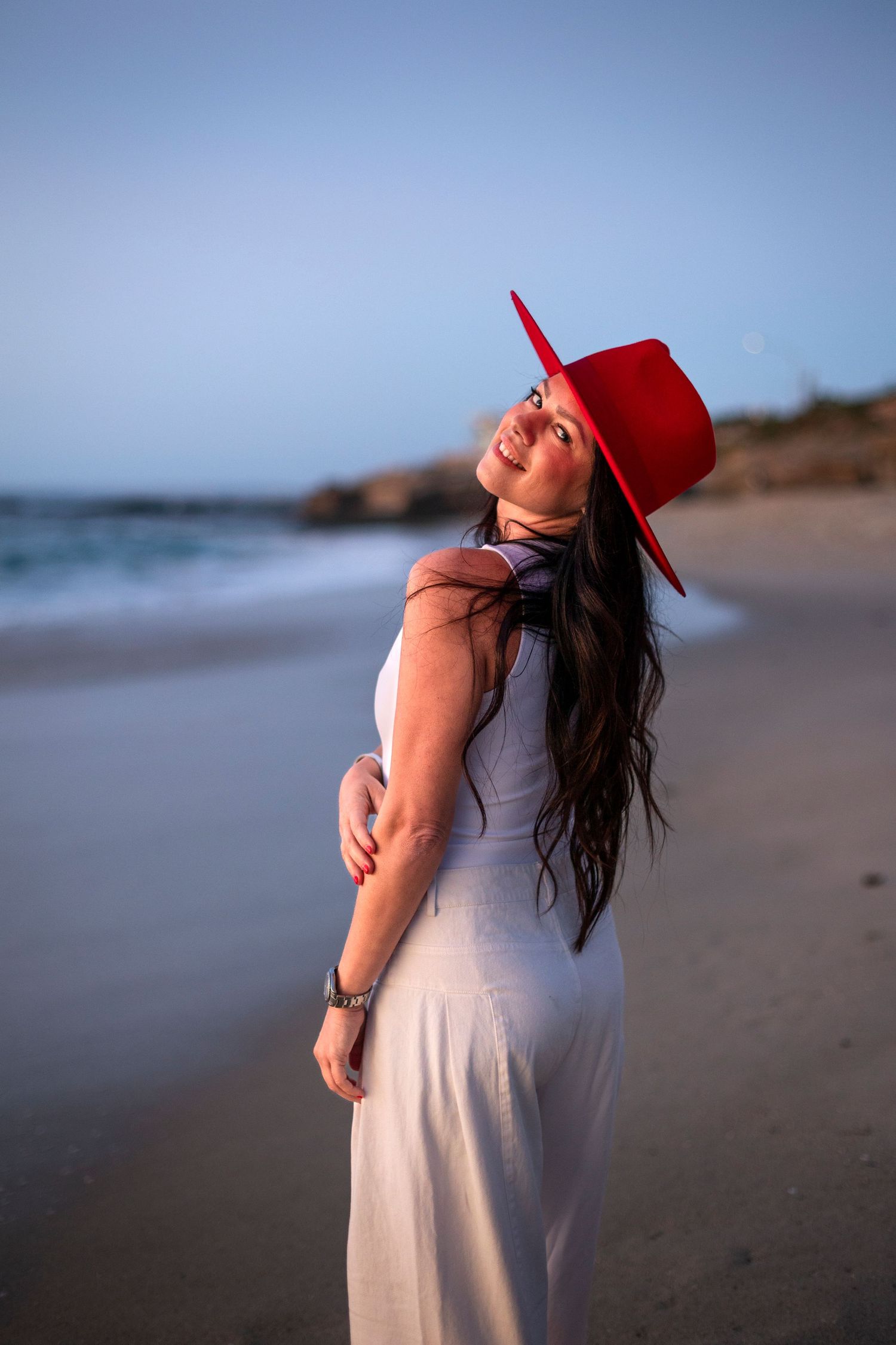 Woman in white dress and red hat smiling on a sandy beach at sunset.