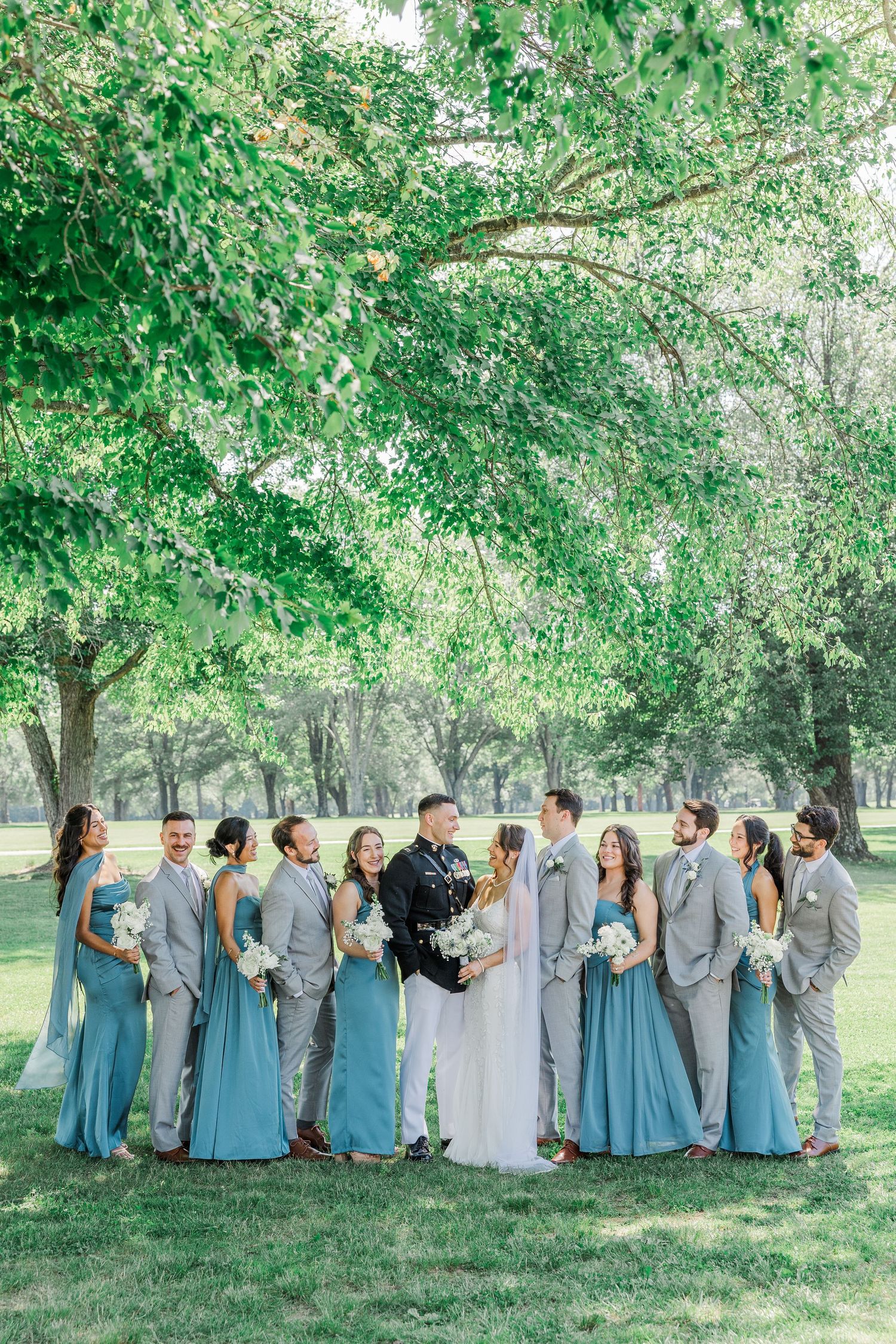 Wedding party poses under lush green trees, bridesmaids in blue gowns and groomsmen in gray suits flanking the bride and groom.
