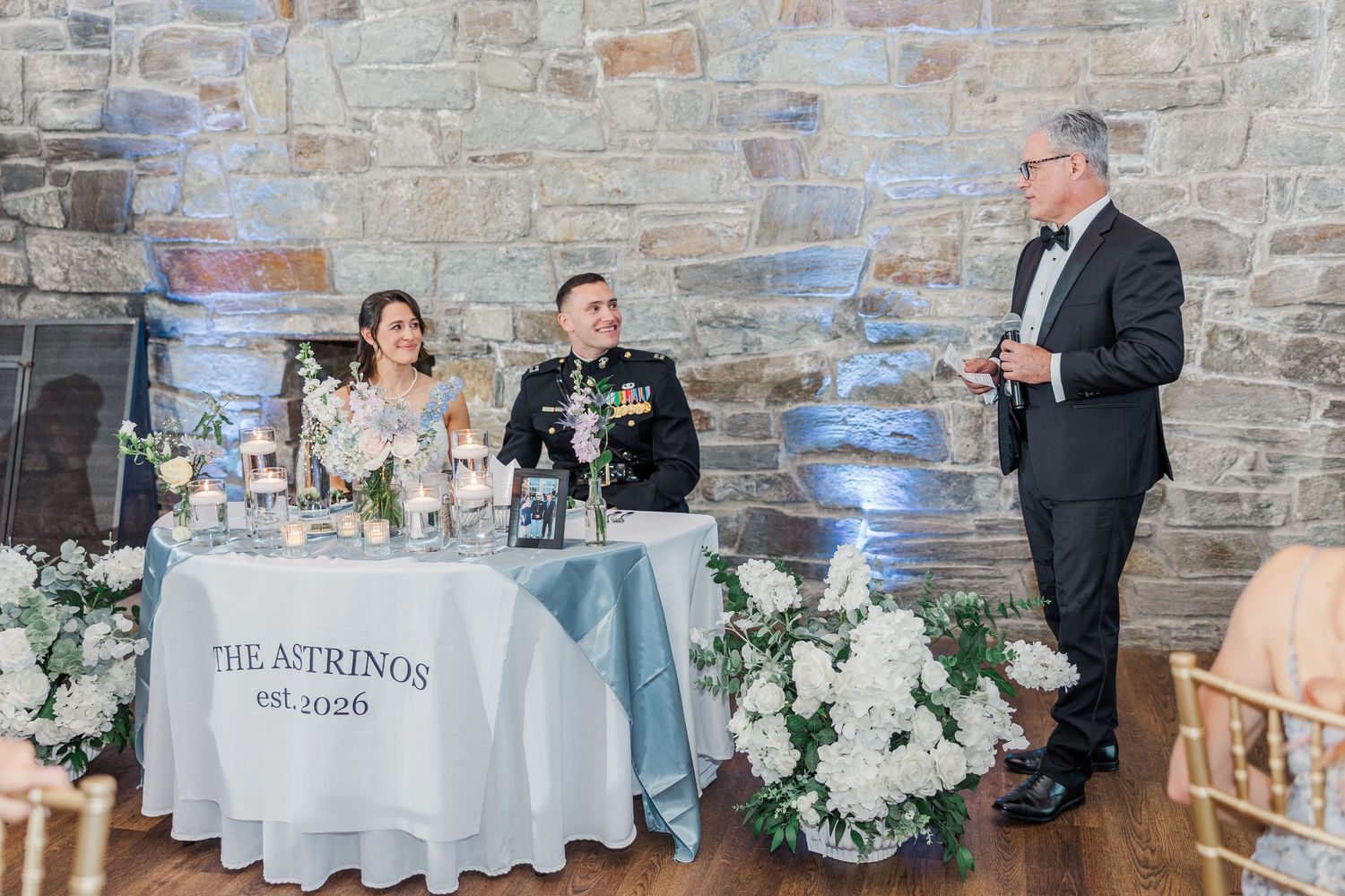 Couple seated at wedding sweetheart table with The Astrinos est 2026 sign while man in tuxedo gives toast.