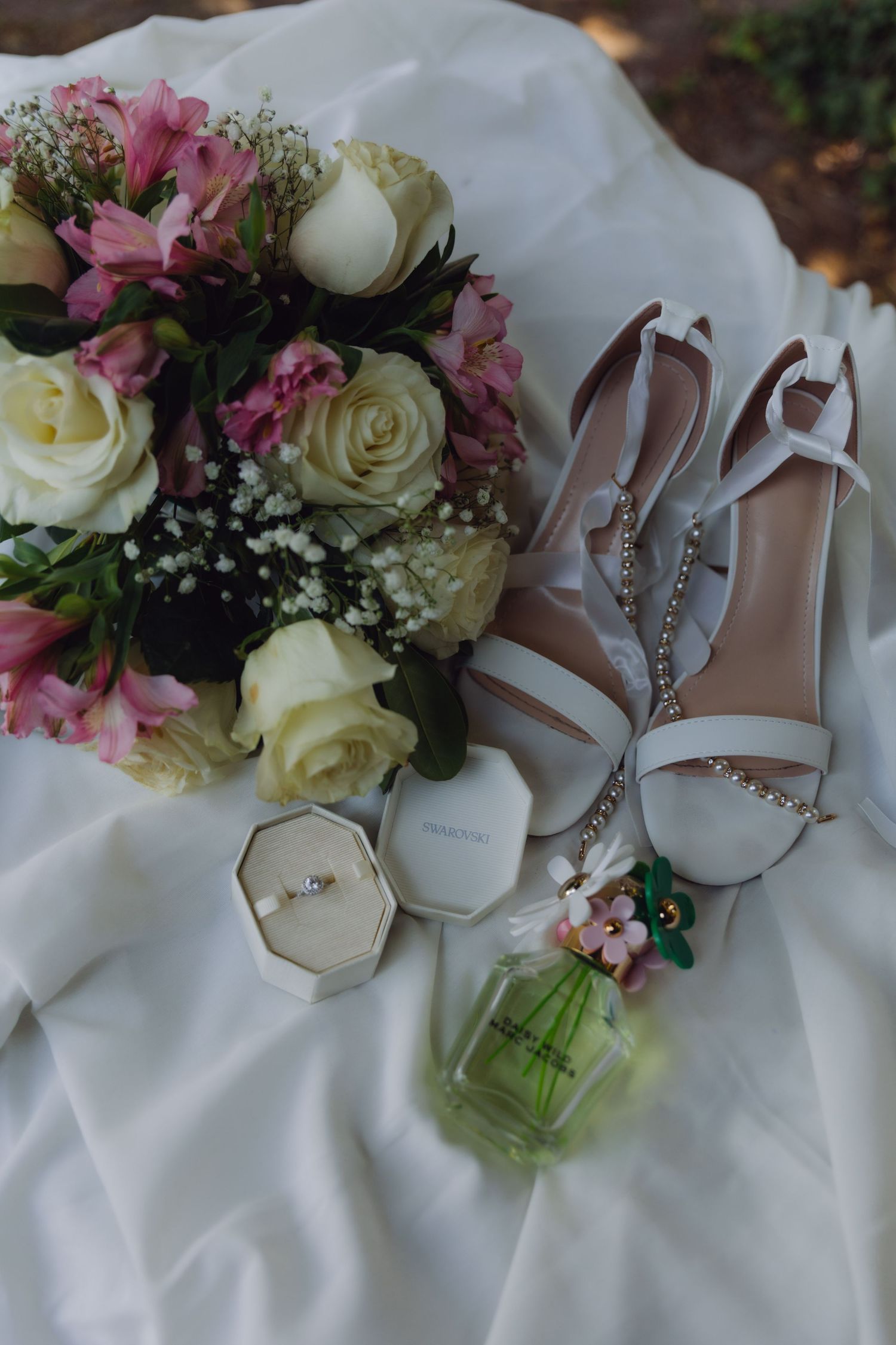 Bridal flat lay with white heels, rose bouquet, ring box, and perfume on white fabric.
