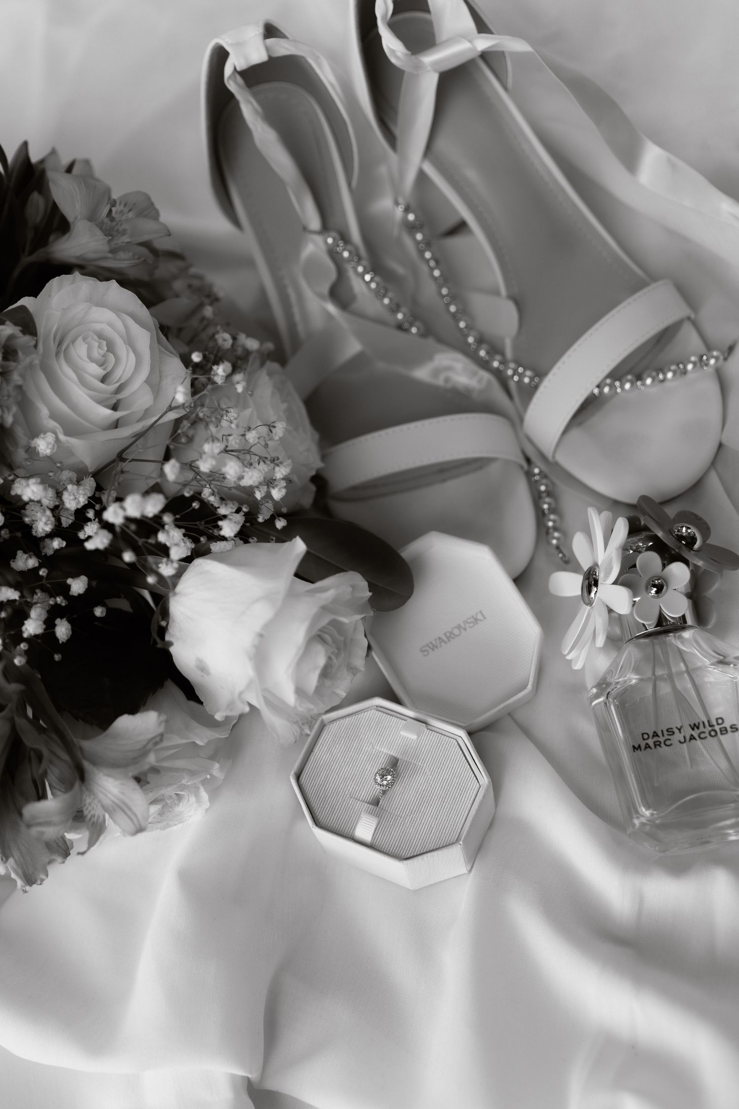 Black and white flat lay of bridal accessories including heels, bouquet, ring box, and Marc Jacobs perfume.