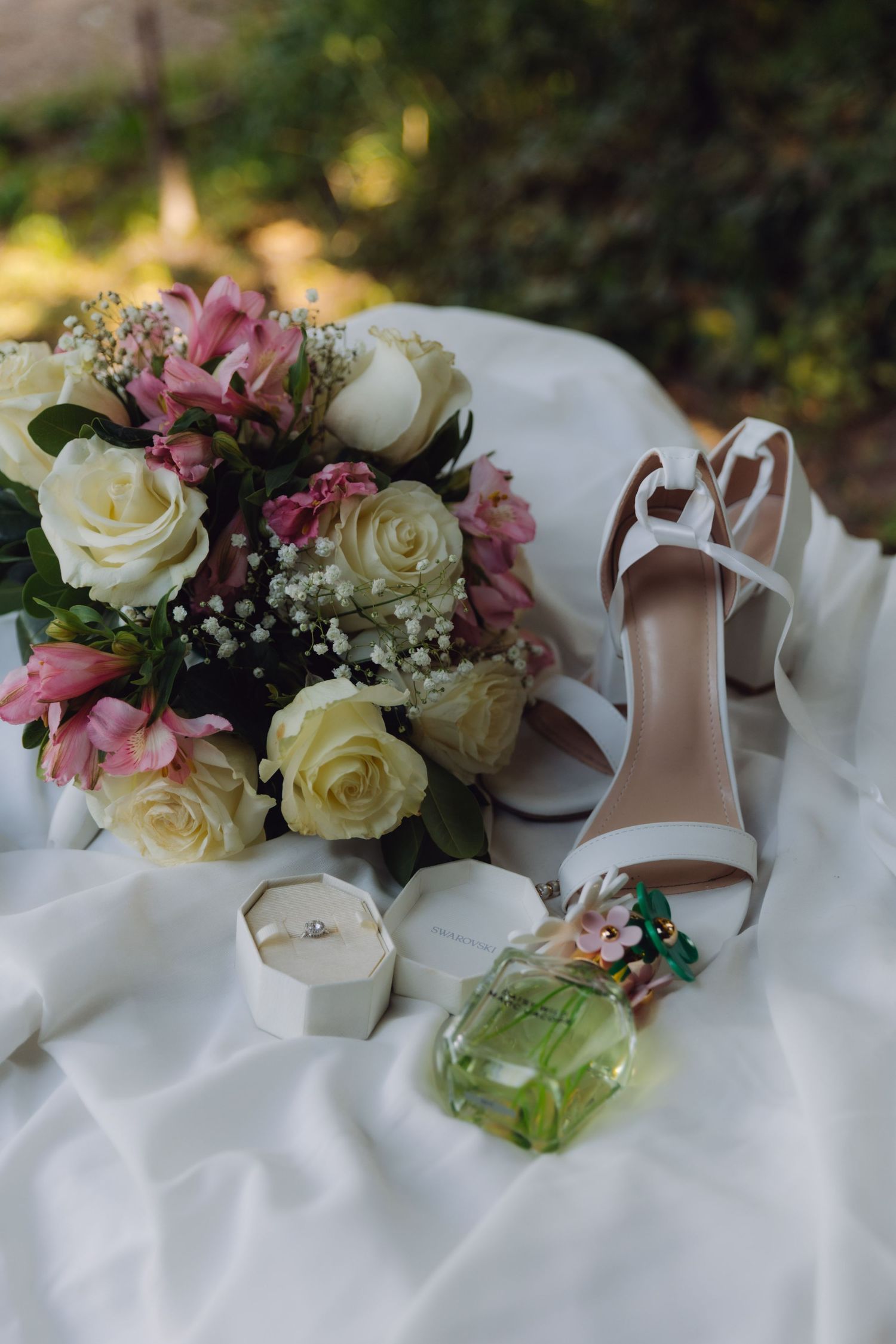 Elegant bridal flat lay featuring white rose bouquet, strappy heels, diamond ring box, and green perfume bottle.