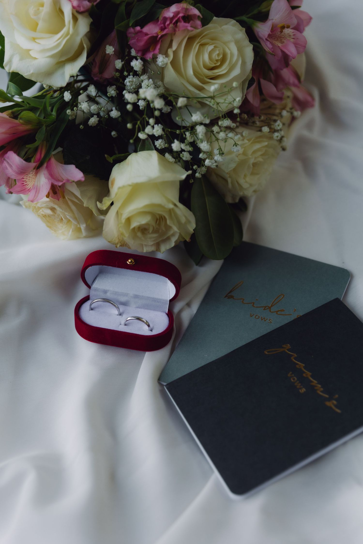 Wedding flat lay with rings in red box, white rose bouquet, and bride and groom journals.