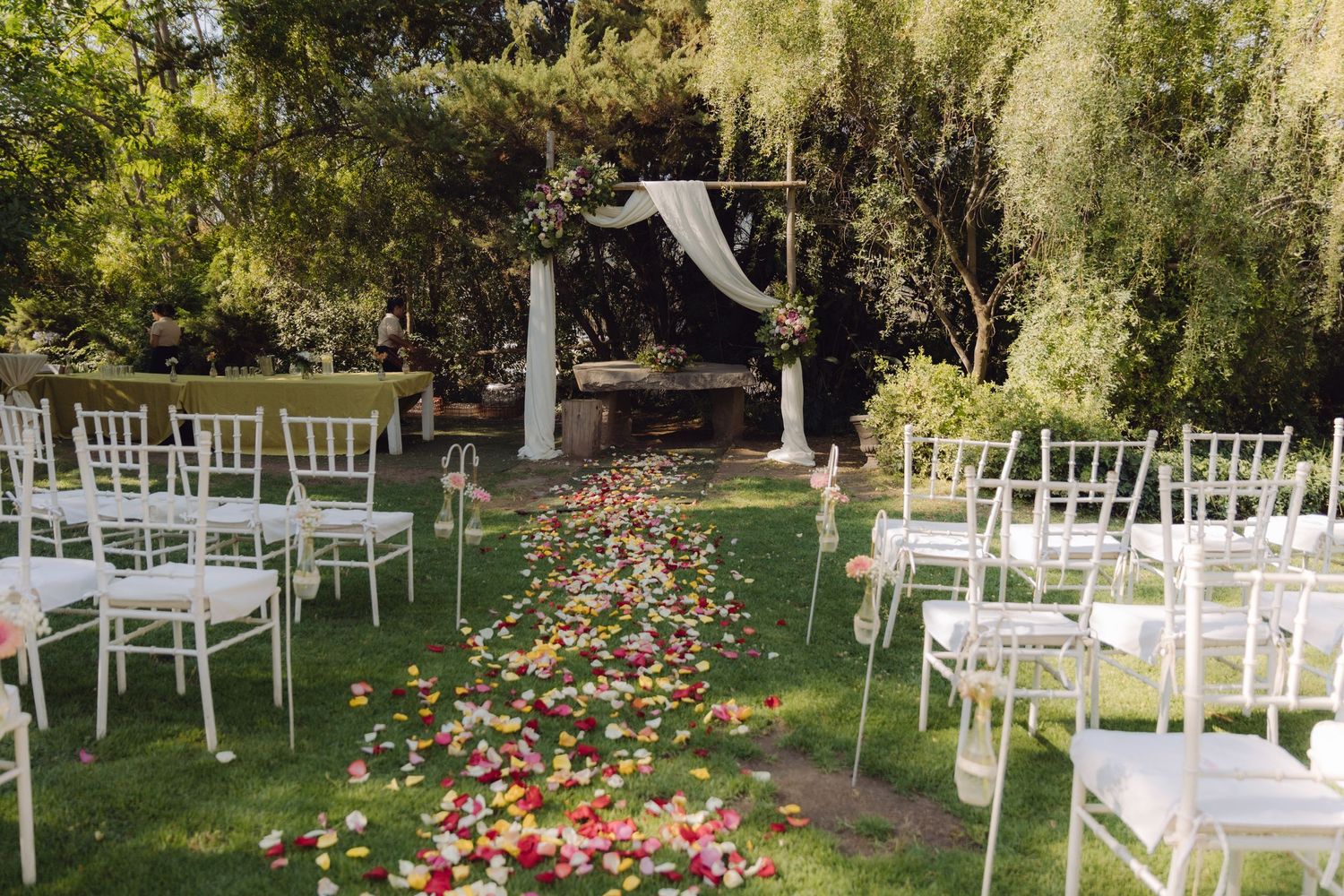 Outdoor wedding ceremony setup with white chairs, floral petal aisle, and draped arch in garden.
