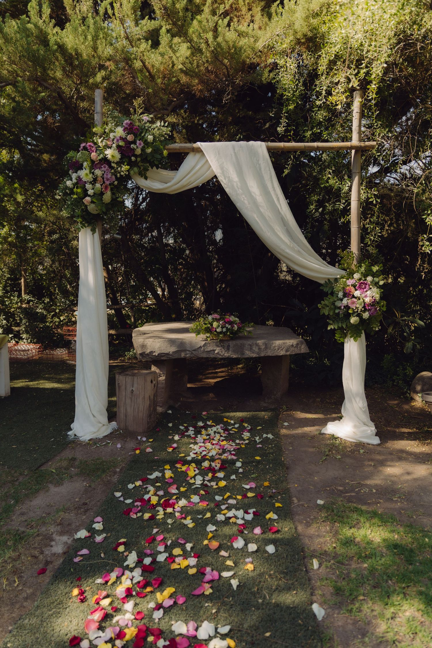 Rustic wooden wedding arch draped in white fabric with floral arrangements and colorful petal aisle.