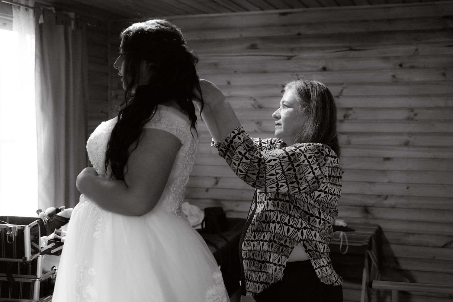 Black and white photo of bride in wedding dress being helped by woman in log cabin.