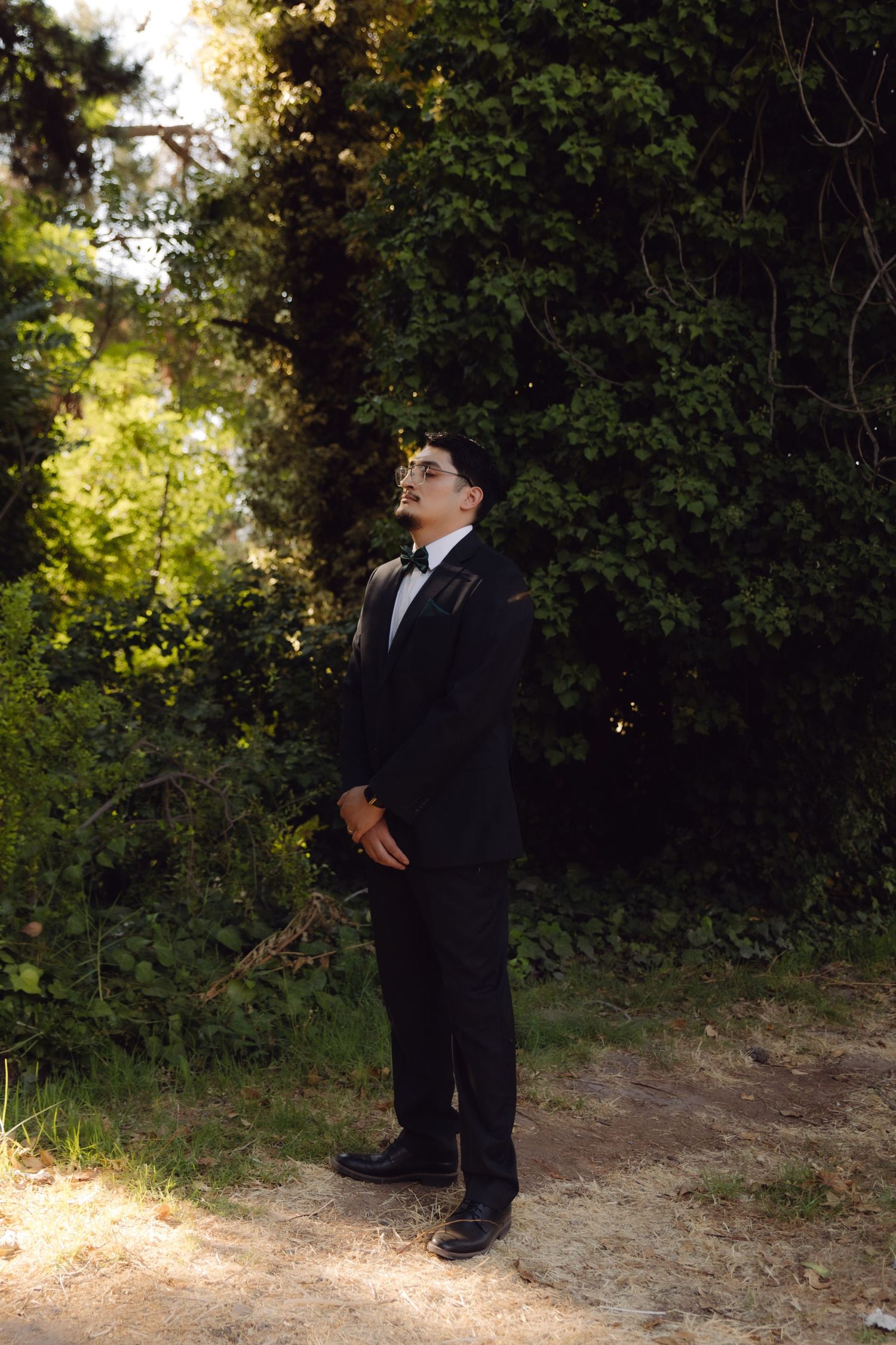 Groom in black tuxedo with bow tie standing outdoors surrounded by lush green trees.