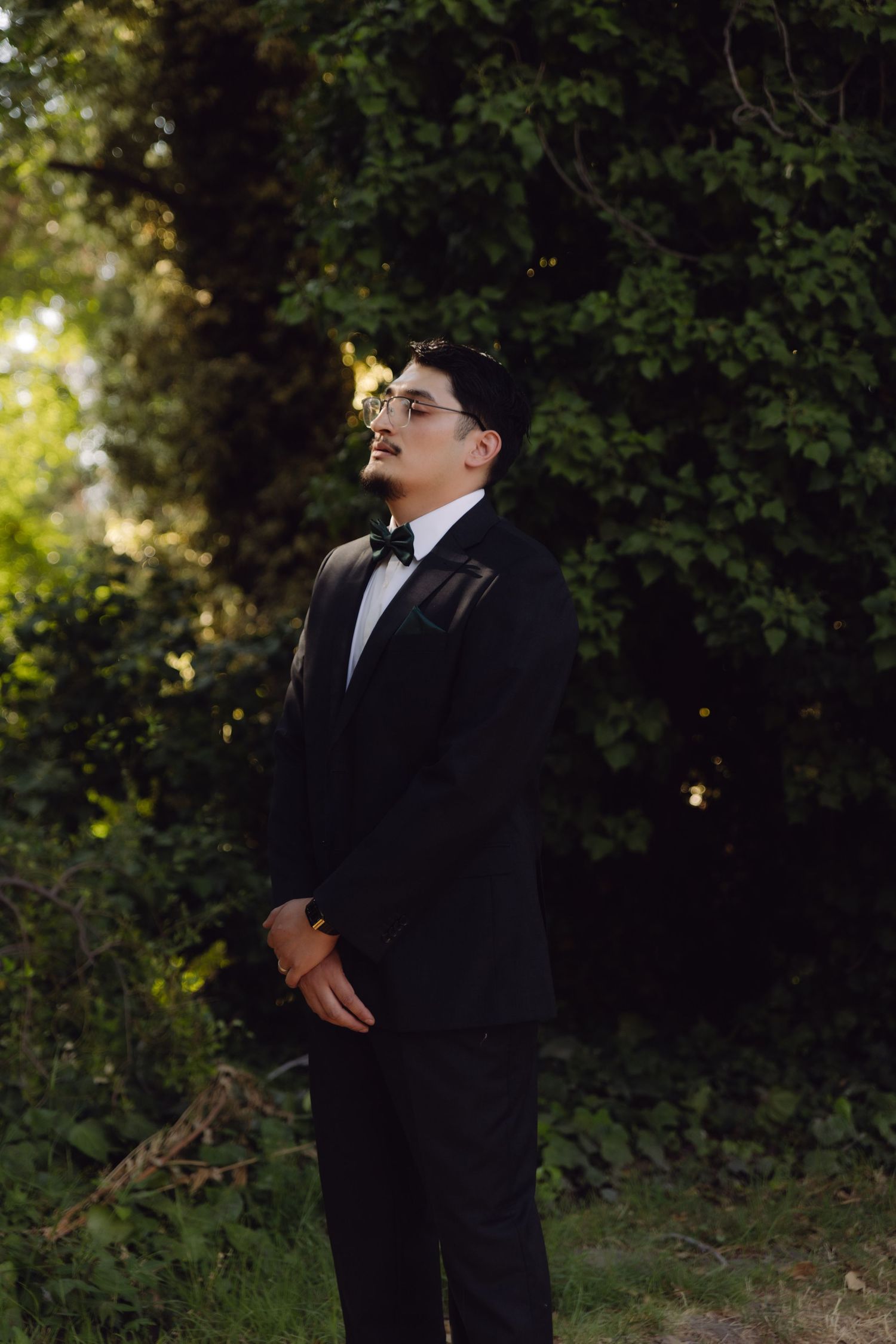 Groom wearing black tuxedo and bow tie looking upward outdoors with green foliage background.