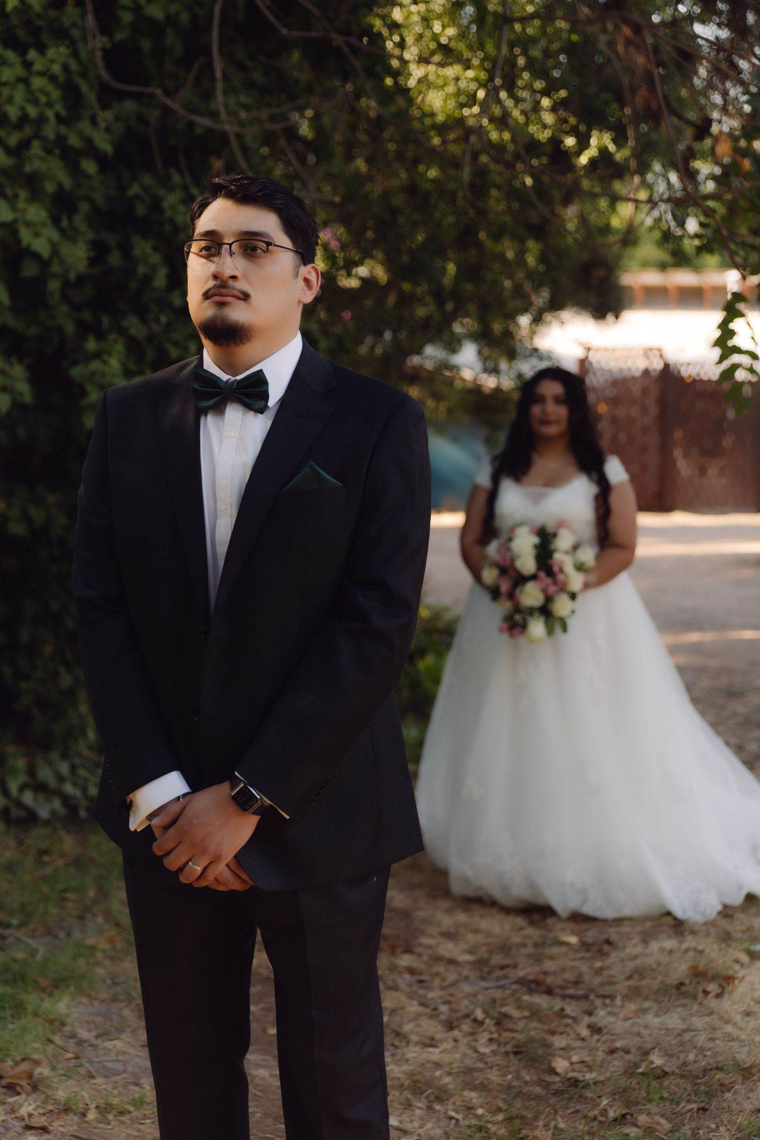 Groom in black tuxedo stands in foreground while bride in white gown holds bouquet in background outdoors.