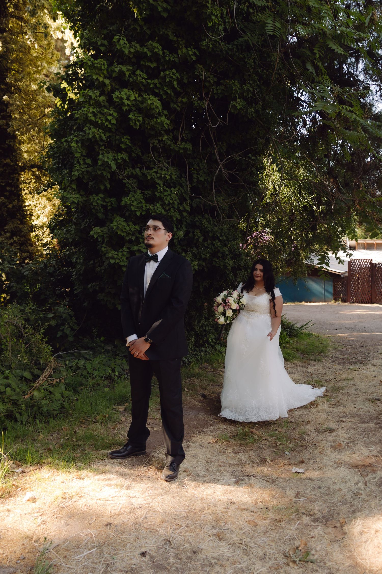 Wedding couple outdoors with groom in tuxedo standing ahead of bride in white gown holding pink bouquet.