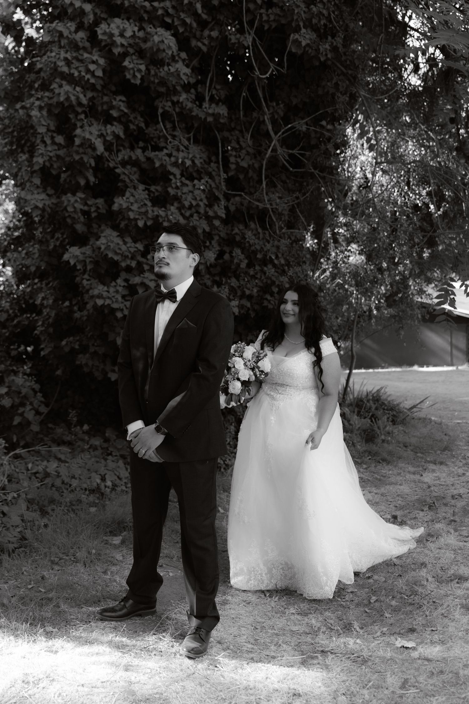 Black and white photo of bride and groom standing apart outdoors beneath lush green trees on wedding day.