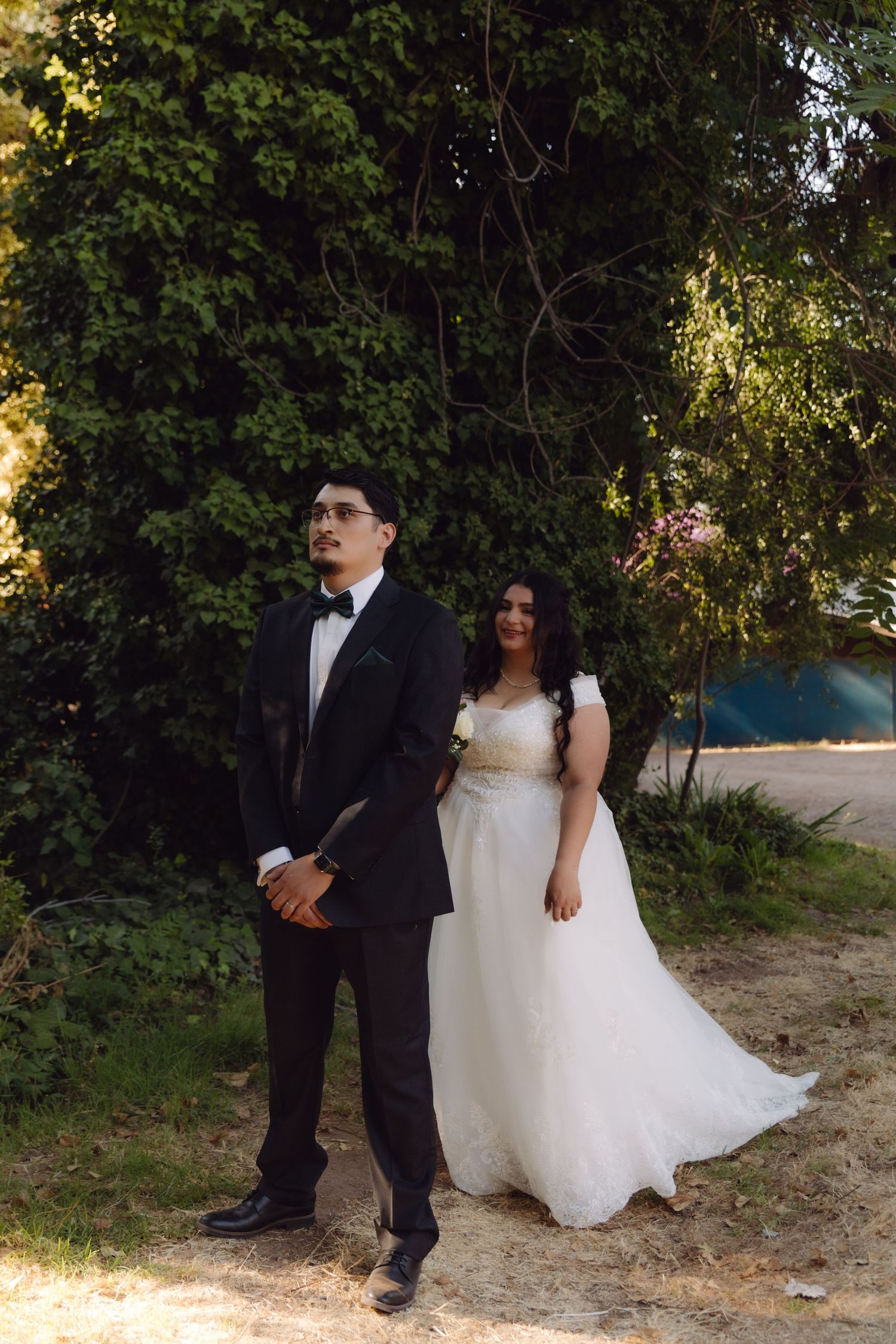 Bride in white off-shoulder gown and groom in black tuxedo pose together in sunny outdoor garden setting.