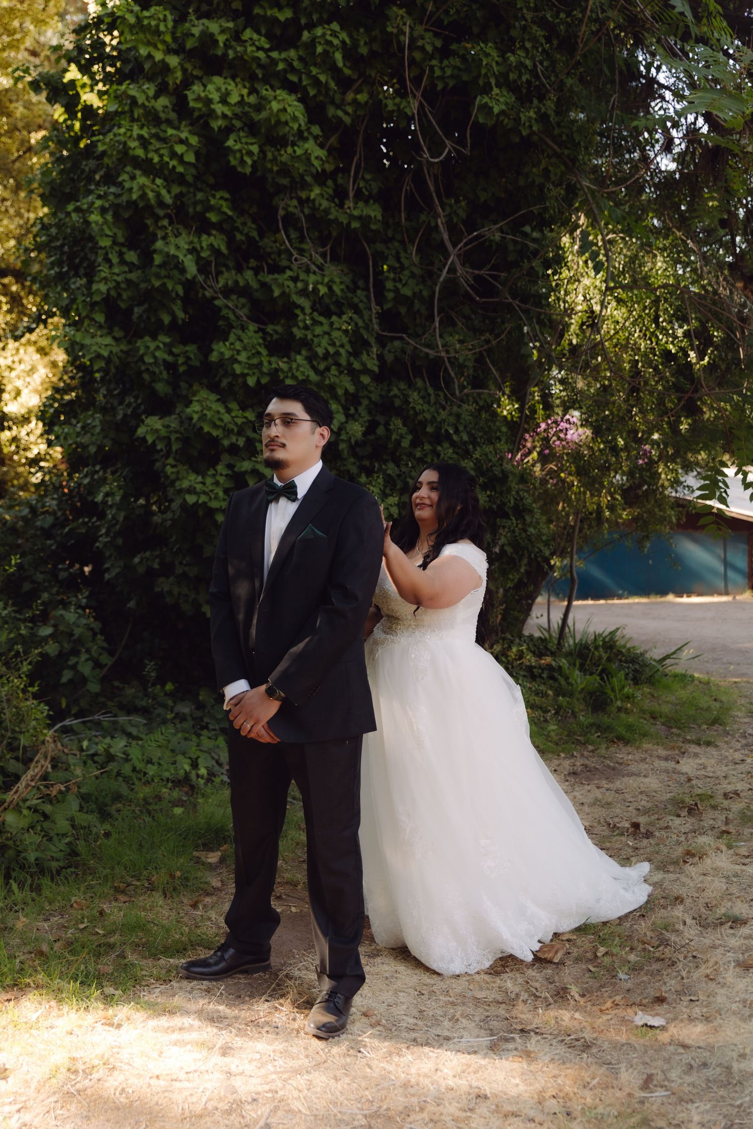 Bride in white wedding gown gently touches grooms shoulder as he stands facing away in black tuxedo outdoors.