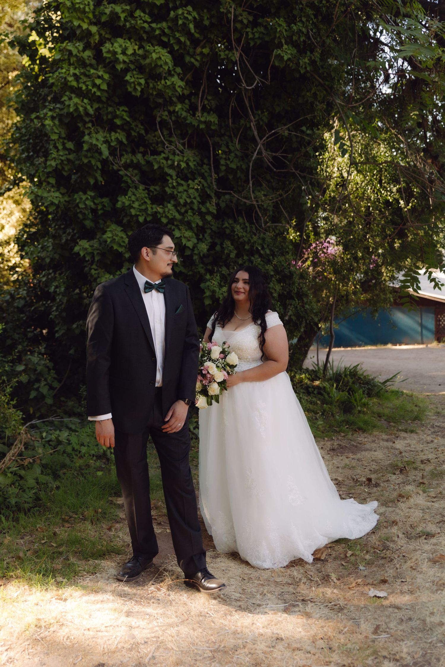 Bride and groom share a romantic glance while standing together outdoors surrounded by green trees on wedding day.