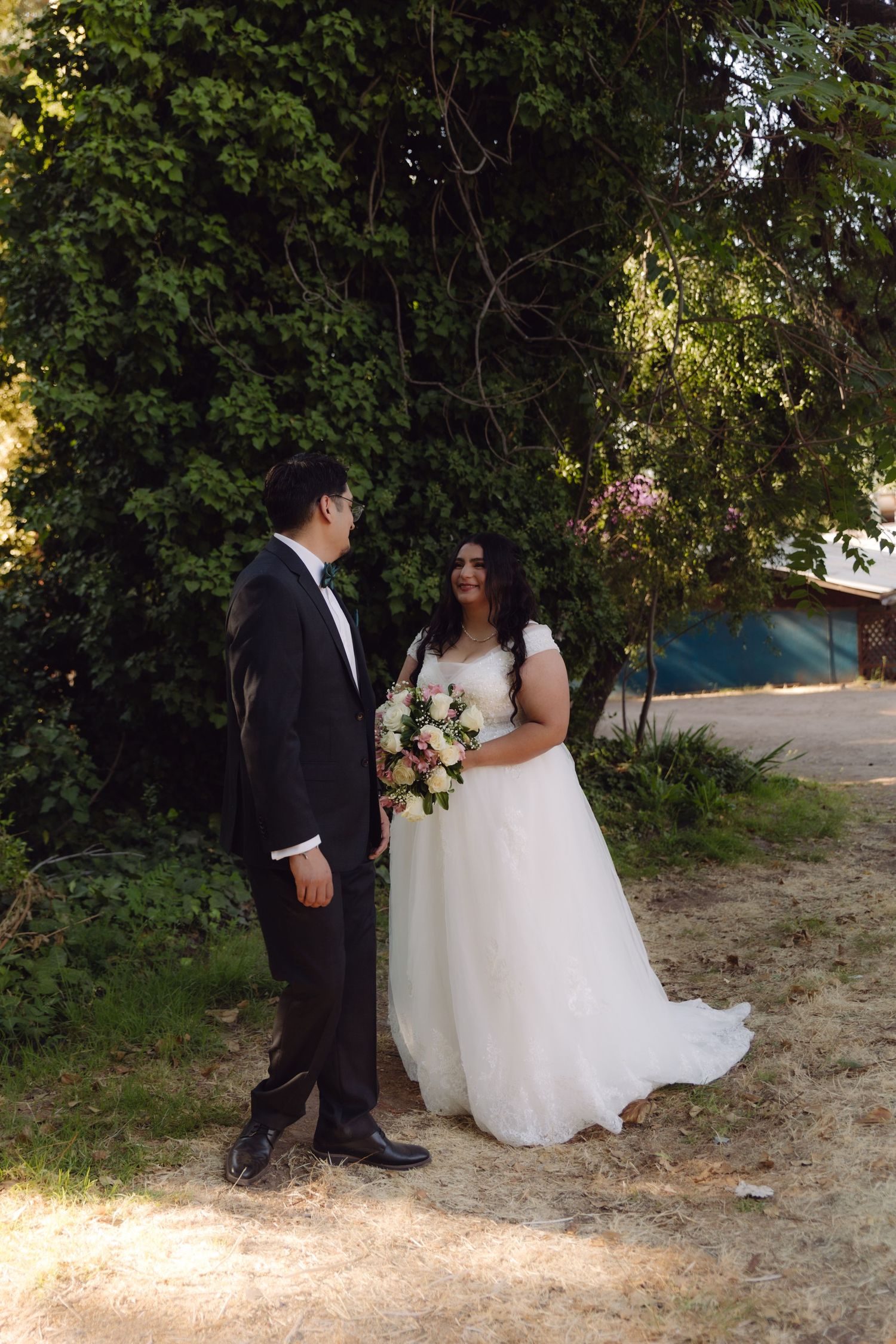 Bride smiles at groom while holding pink and white bouquet as couple stands facing each other outdoors.