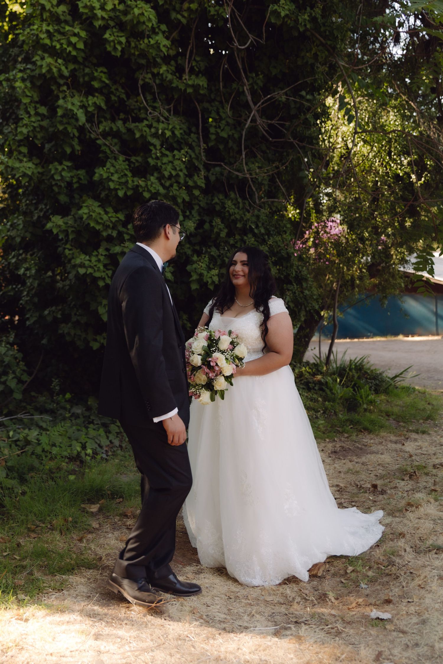 Bride in white gown smiles at groom in black tuxedo while holding bouquet in a sunlit outdoor setting.