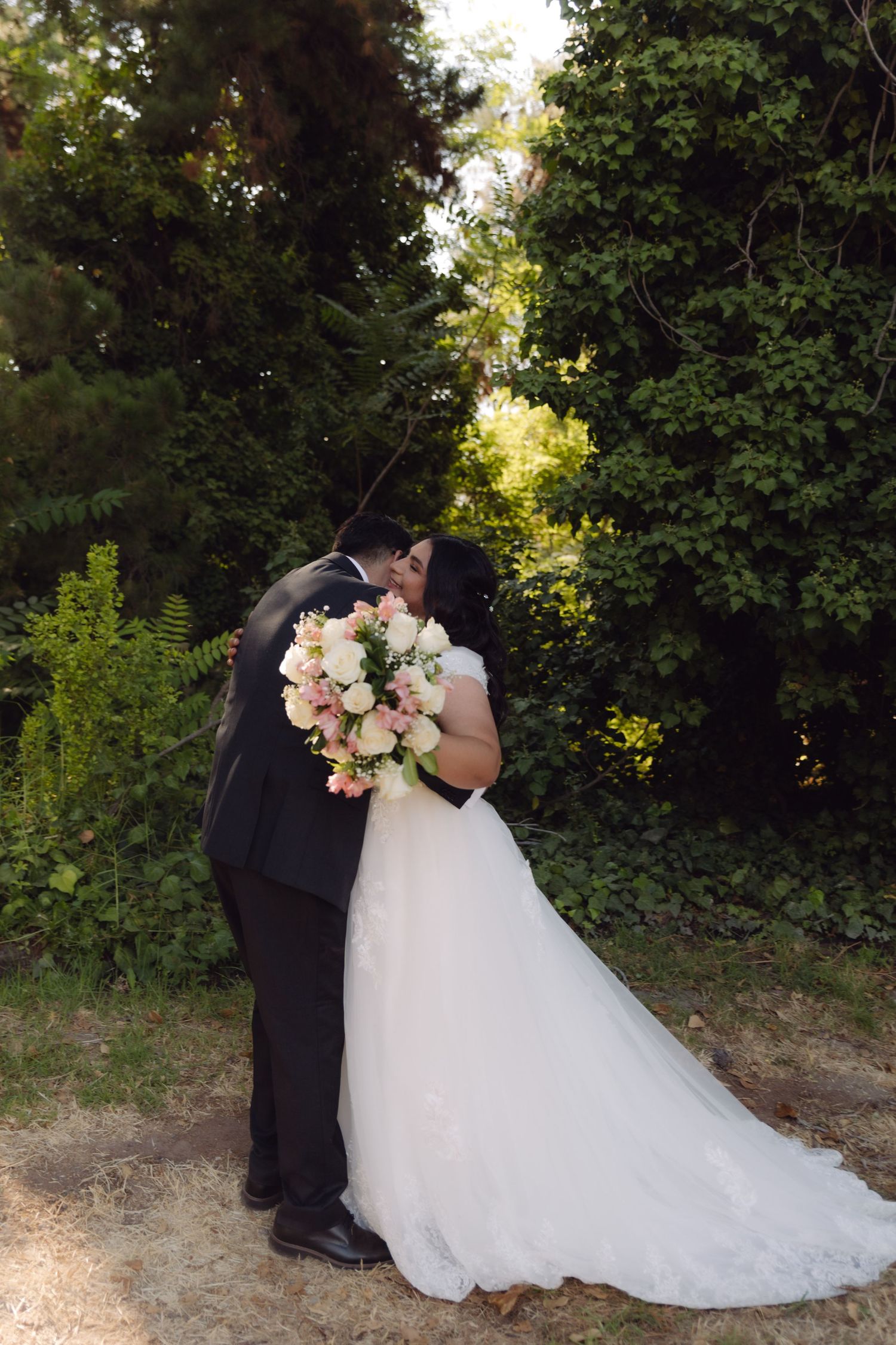 Bride and groom share a kiss outdoors while bride holds a pink and white floral bouquet on wedding day.