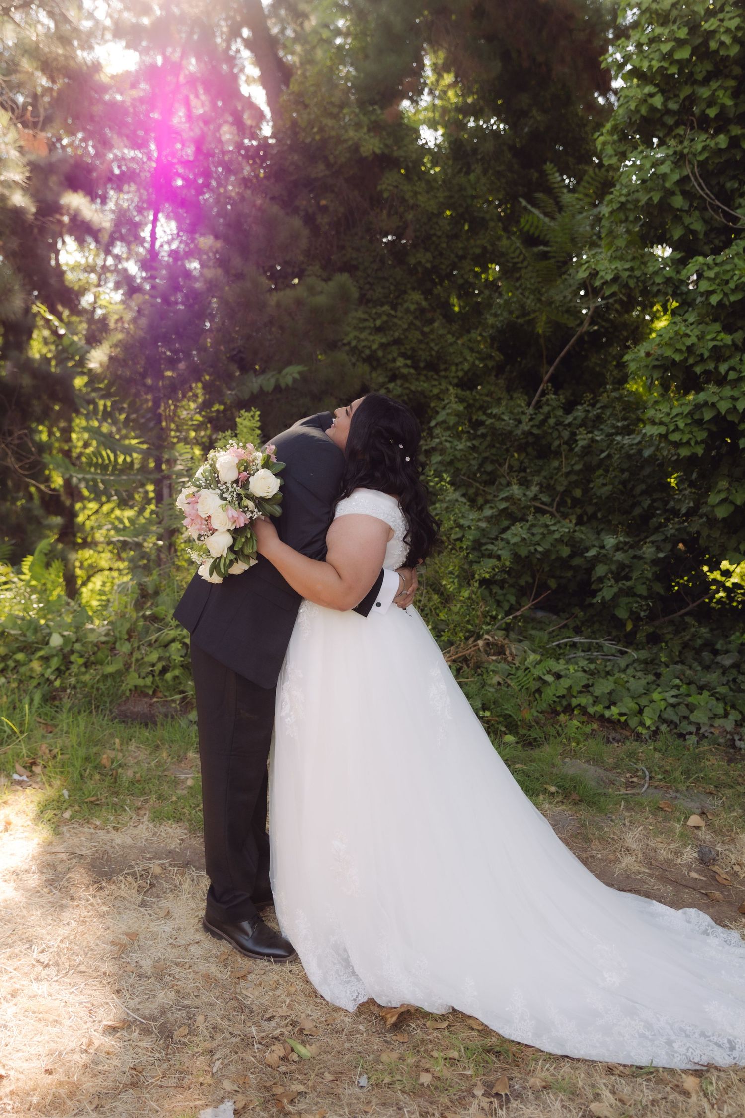 Bride and groom embrace and kiss in sunlit outdoor garden with lens flare and lush green trees behind them.