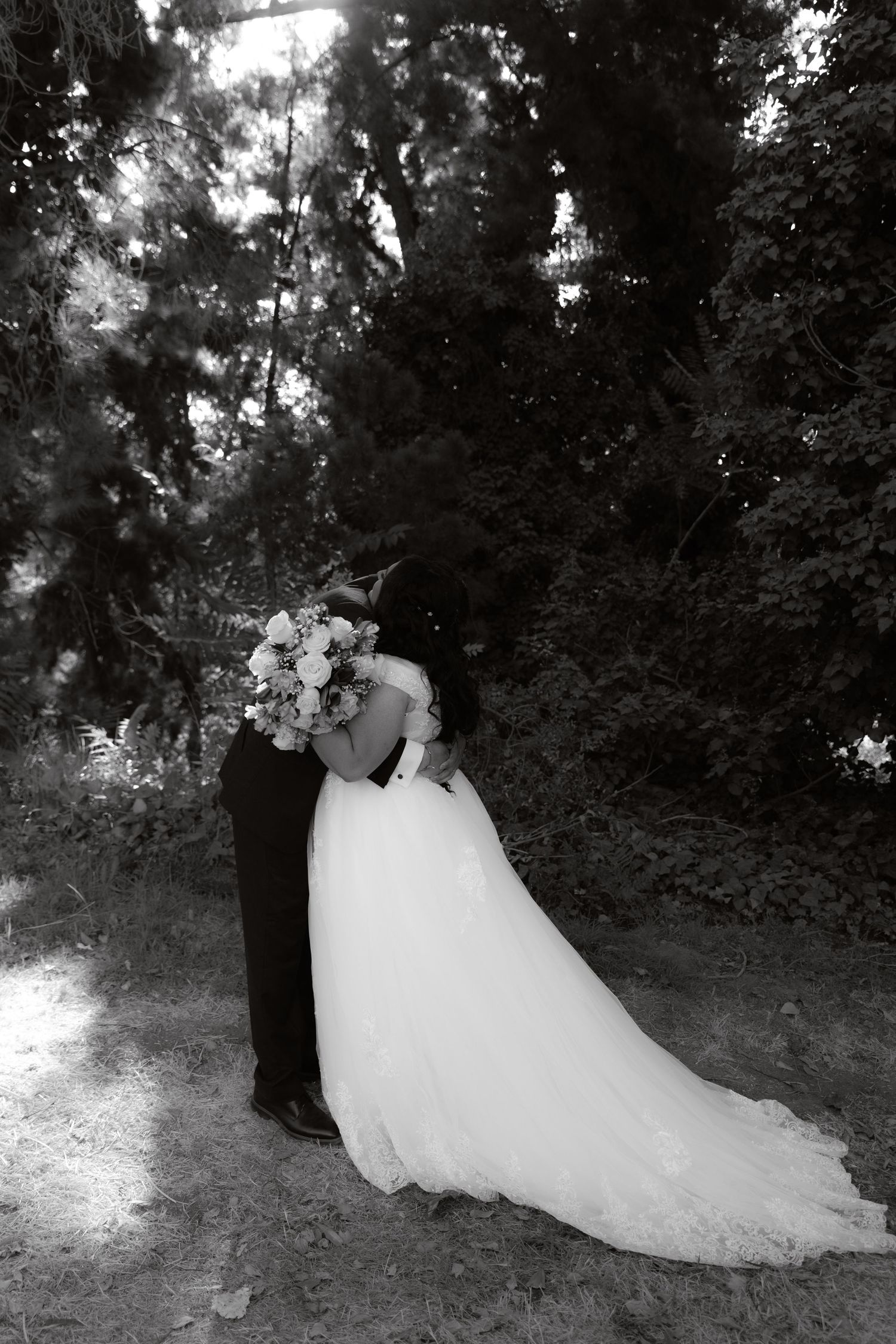 Black and white photo of bride and groom embracing in a lush forest setting on their wedding day.