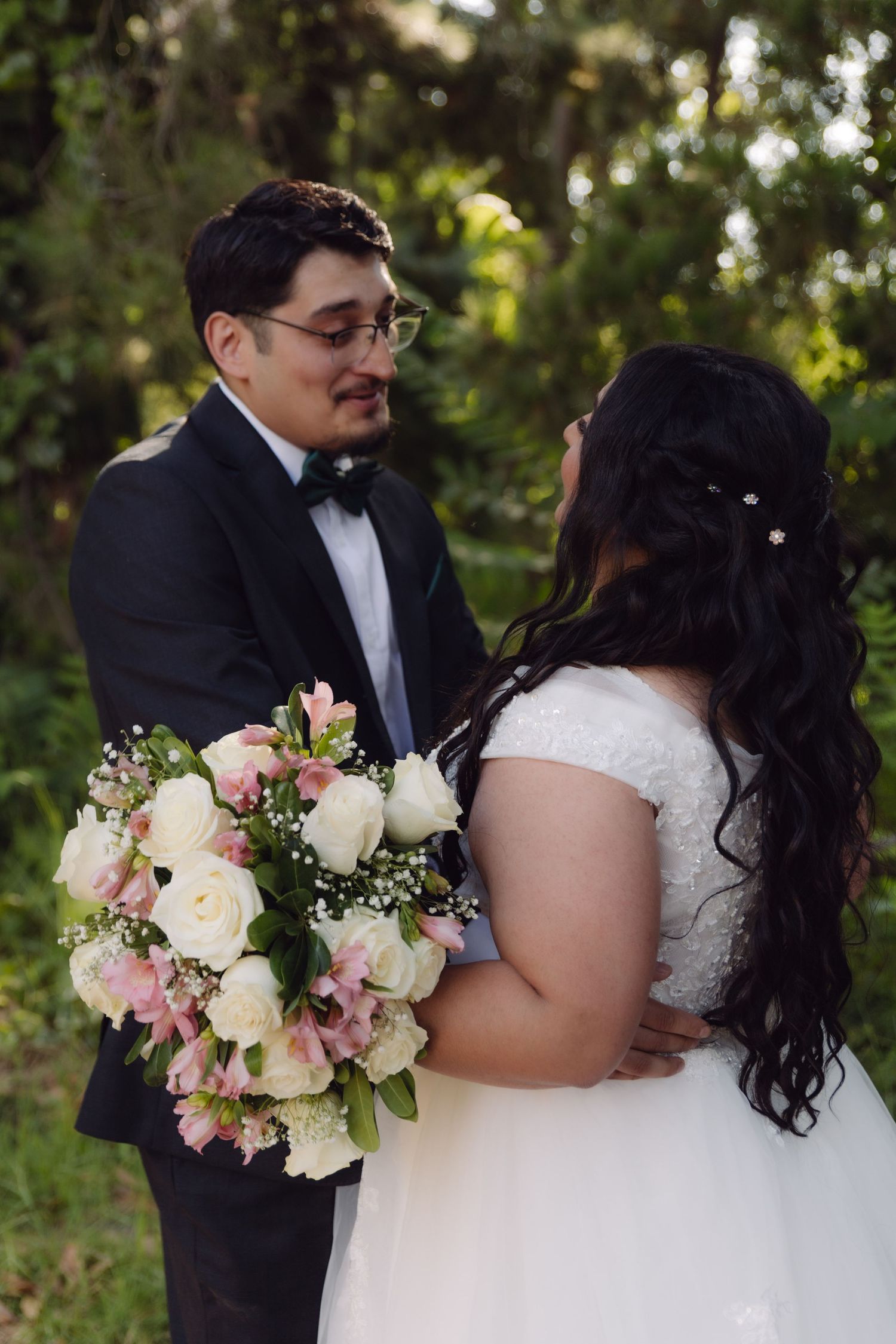 Groom in black tuxedo smiles at bride holding pink and white rose bouquet outdoors.