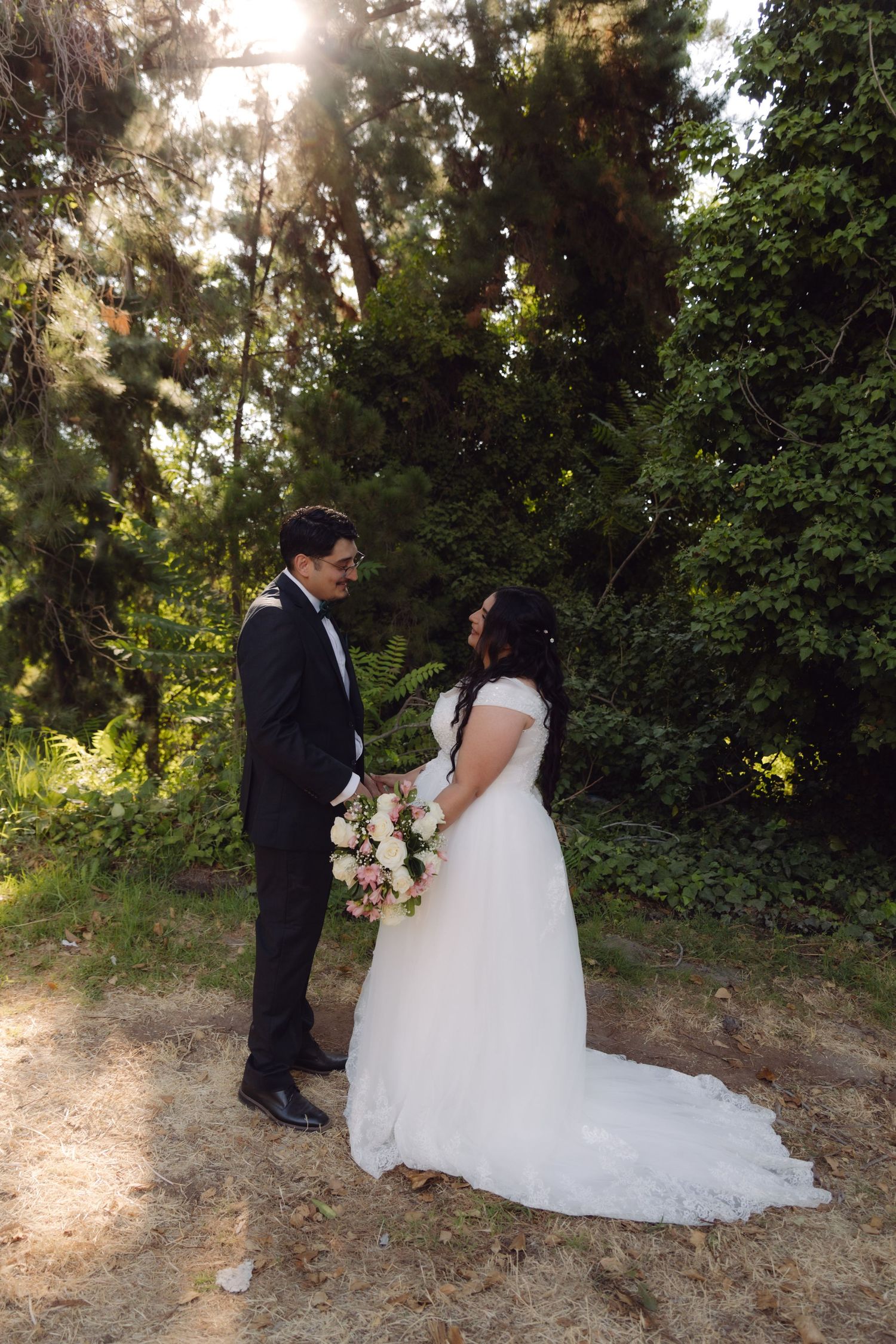 Bride and groom standing face to face in a sunlit forest clearing on their wedding day.