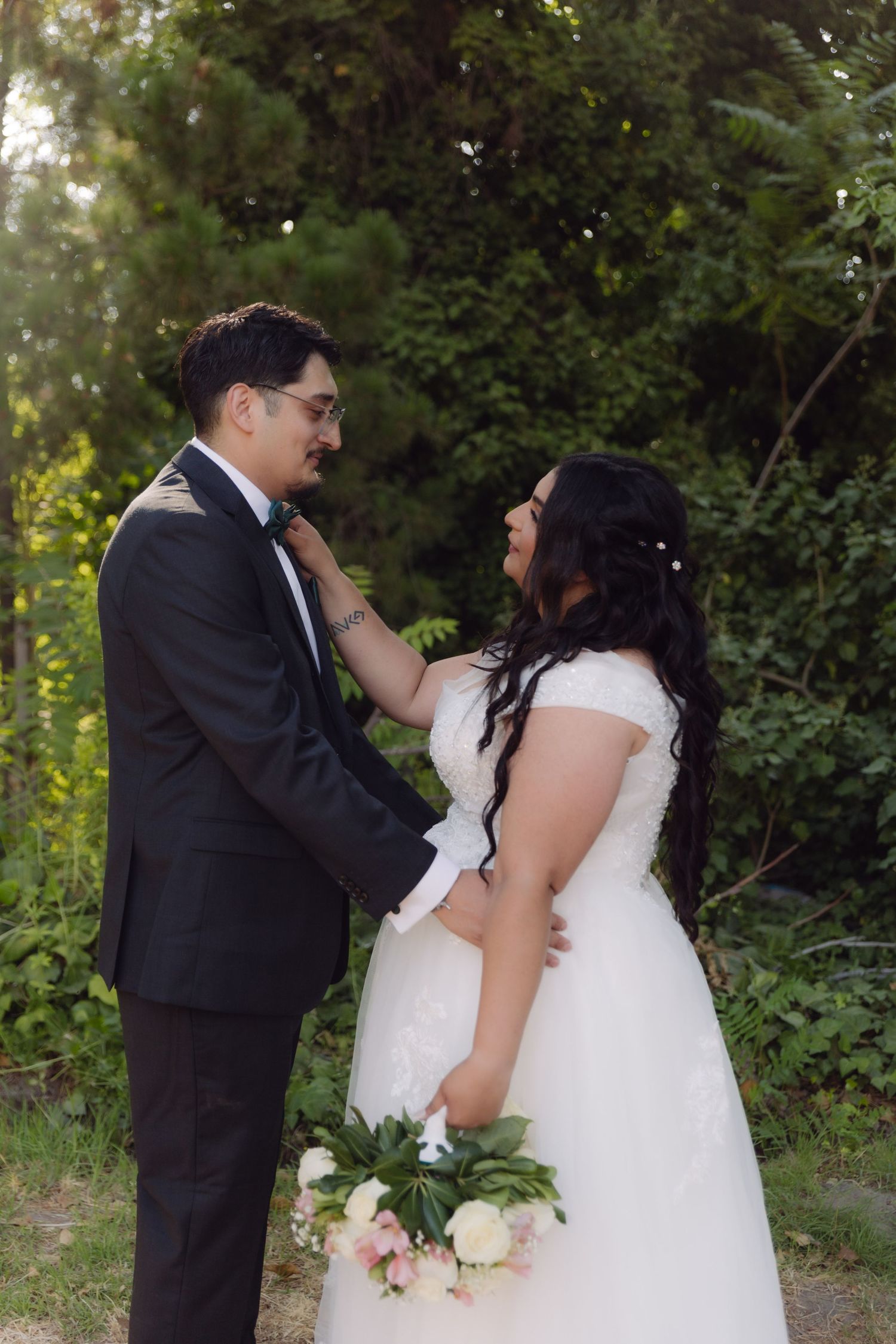 Bride adjusting grooms bow tie while smiling at each other in a green outdoor setting.