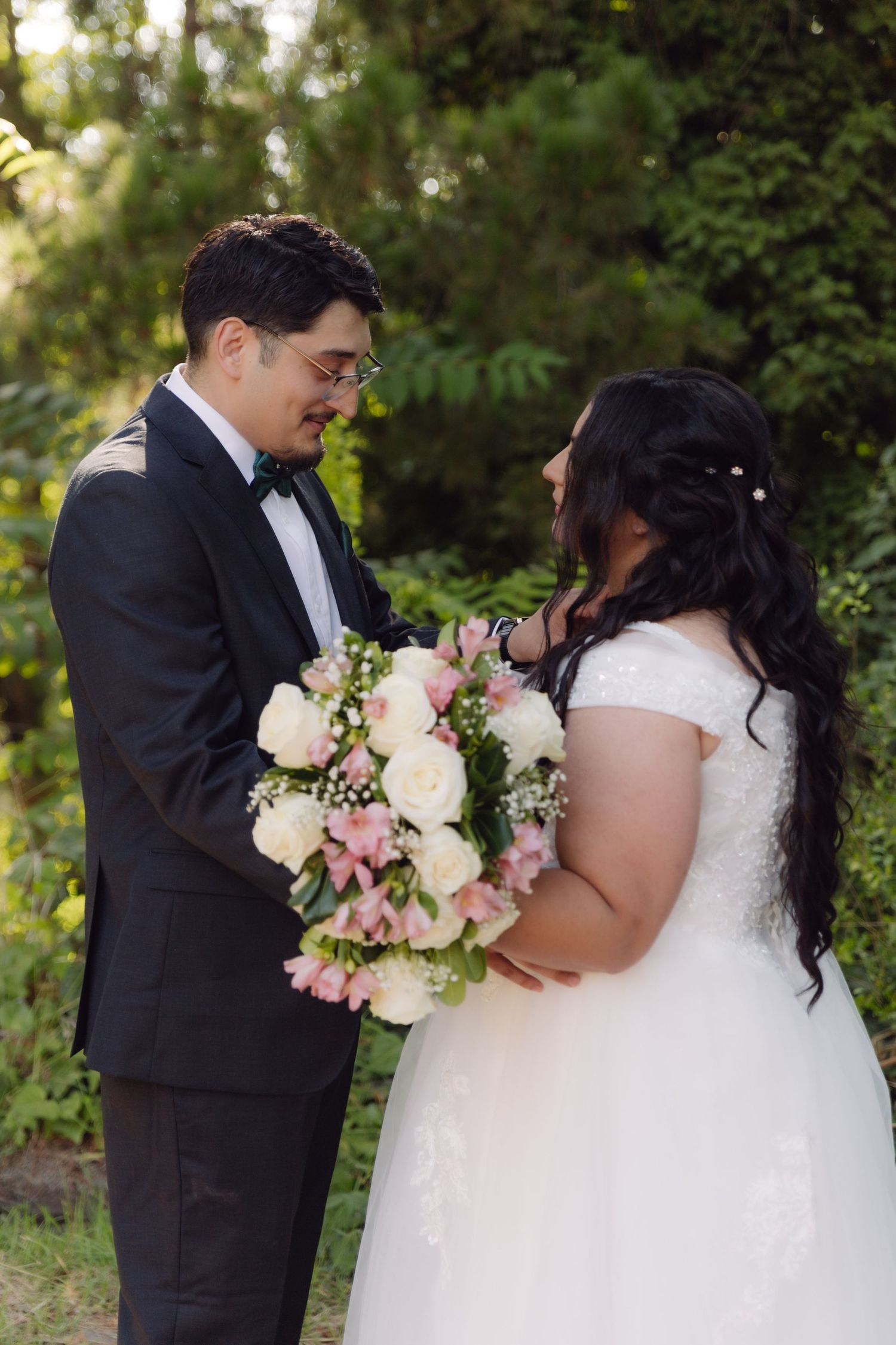 Groom gazing at bride while she holds a pink and white rose bouquet in a garden setting.