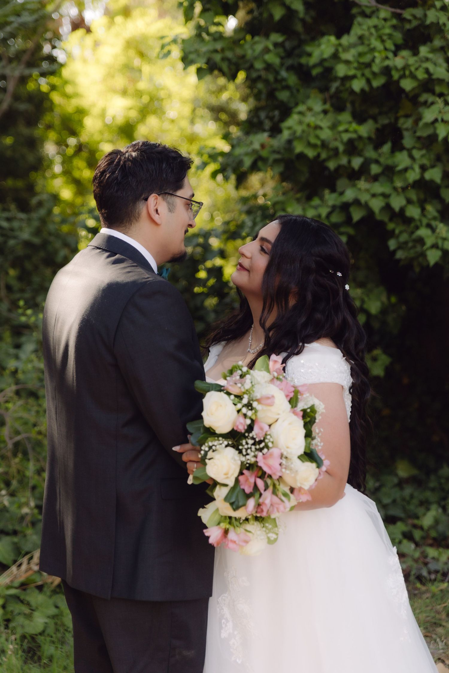 Bride and groom gazing at each other outdoors, bride holding pink and white bouquet in white lace wedding gown.